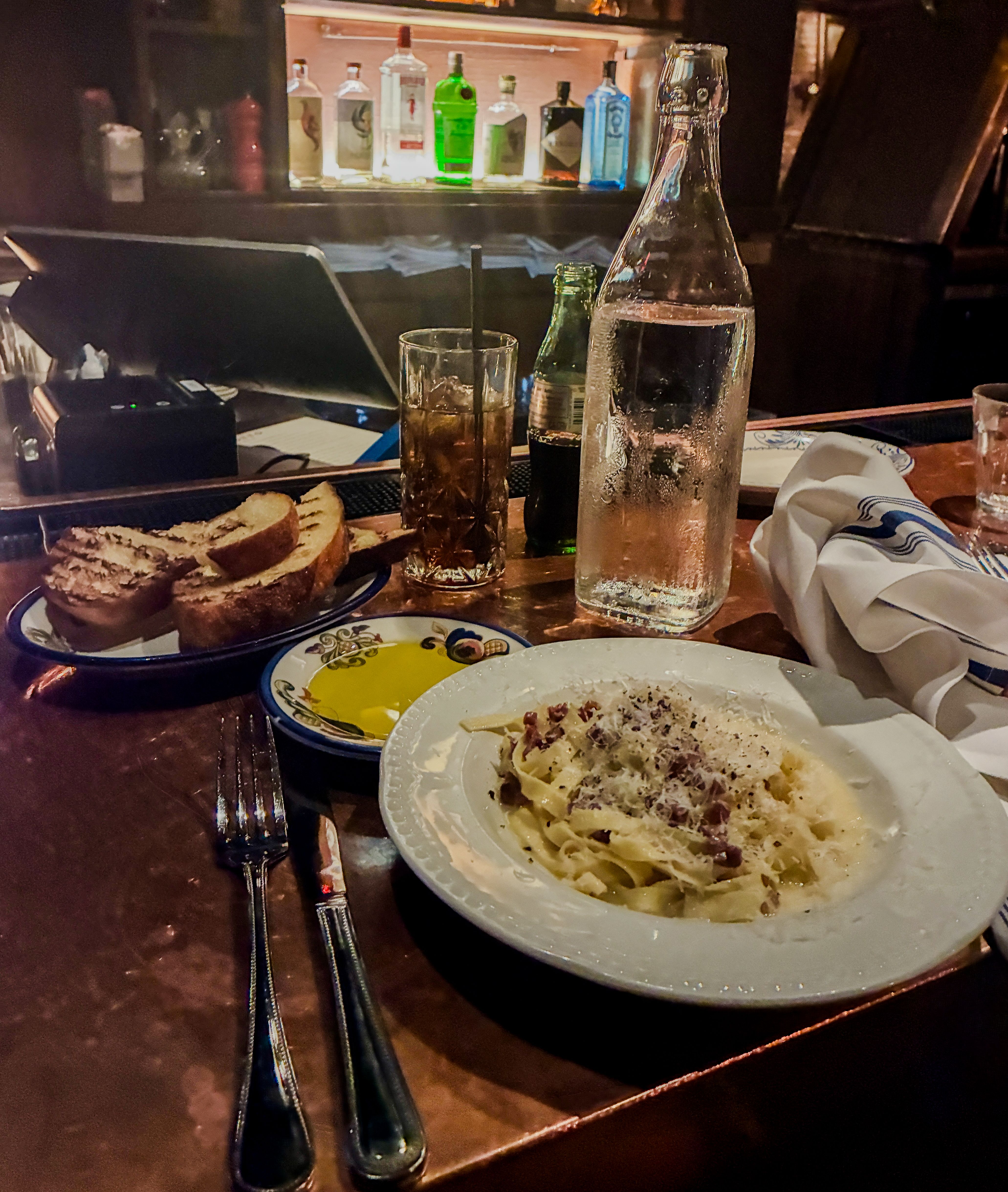 A dim bar table with plates of pasta and bread, a tall glass water bottle, a glass with ice and a straw, forks, and a backlit shelf of colorful liquor bottles.