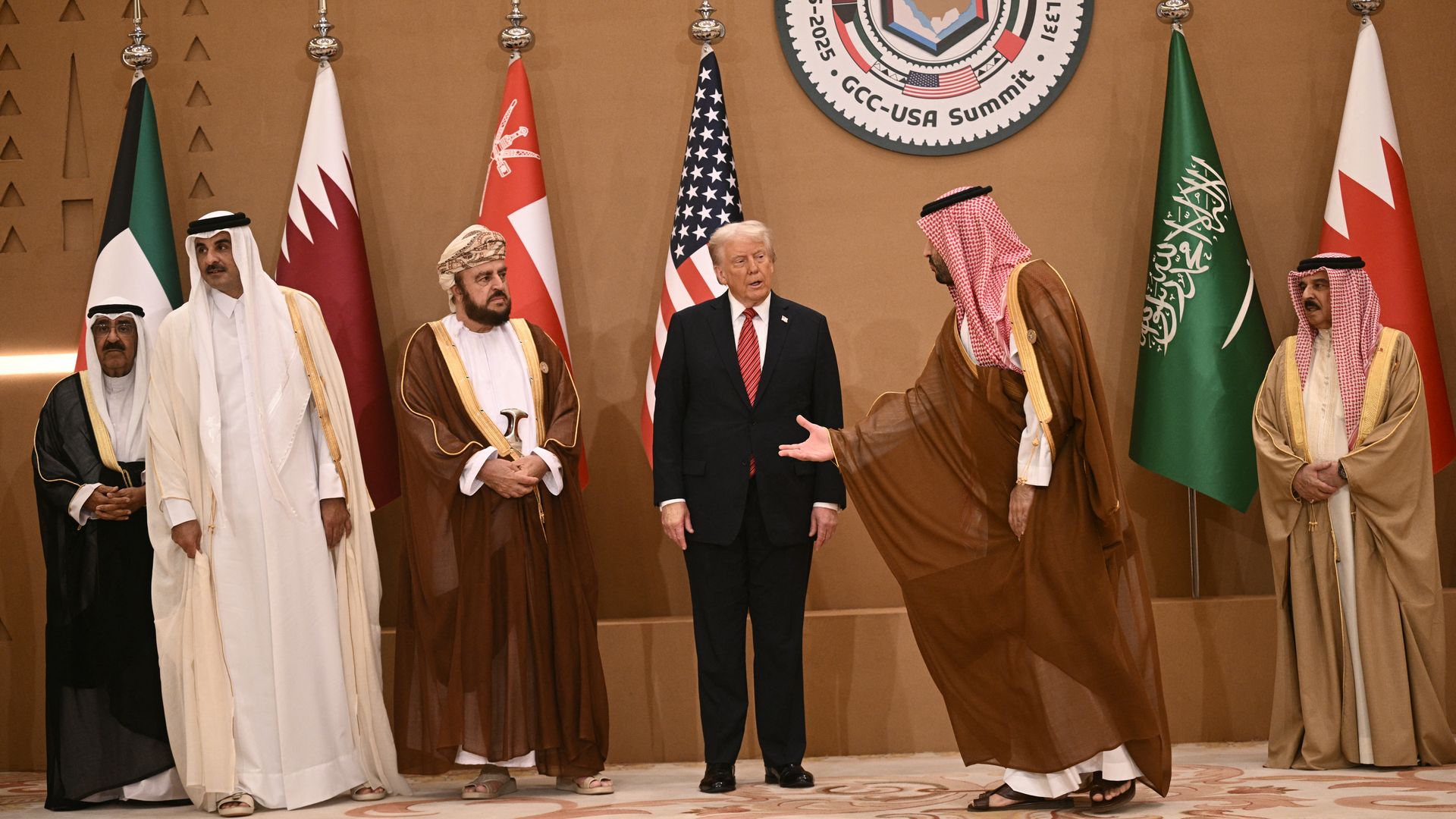 Group of Gulf leaders and U.S. President Donald Trump on a stage at a GCC-USA Summit, flanked by national flags (including U.S. and Saudi) with a large GCC-USA seal behind them.