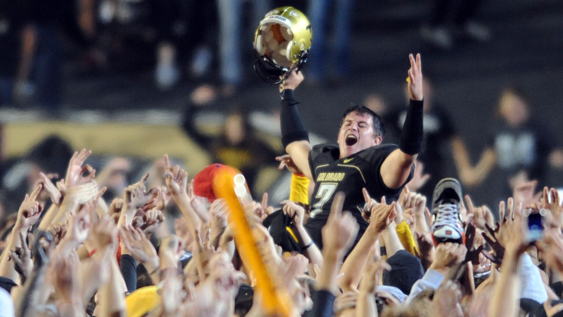 Cody Hawkins wearing black and gold Colorado jersey no. 7 is lifted by cheering crowd, holding helmet high and celebrating enthusiastically at night game.