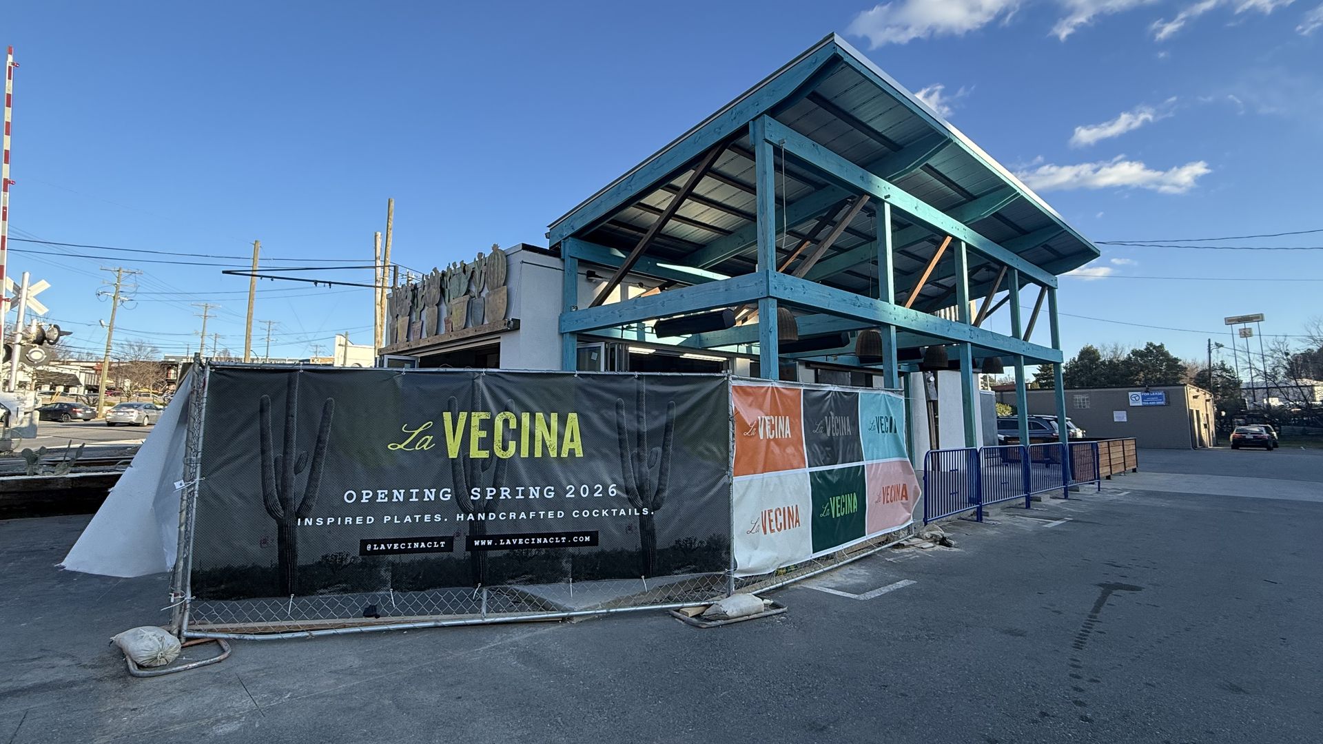 Outdoor construction site for La Vecina restaurant opening Spring 2026, with turquoise wooden framework, banners with cactus images and text about handcrafted cocktails, under a clear blue sky.