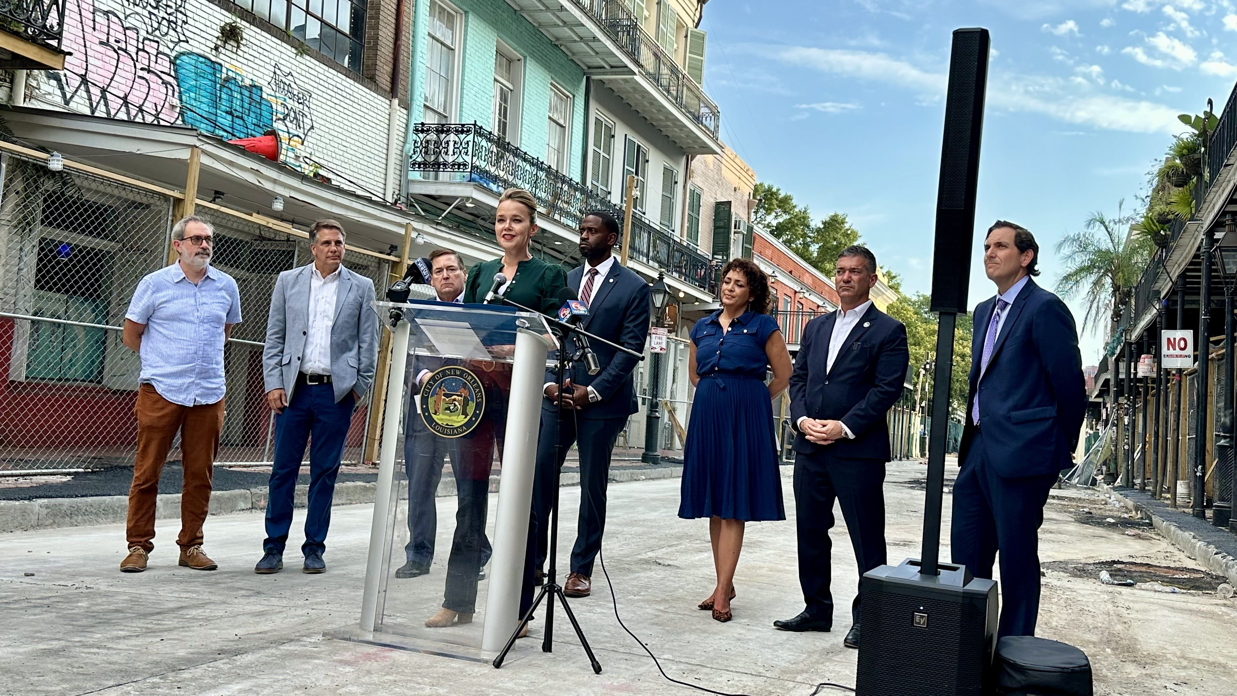 Group of officials at a podium with a city seal on a sunlit street; a woman in green speaks while others in suits listen. Graffiti-covered buildings, balconies, and blue sky behind.