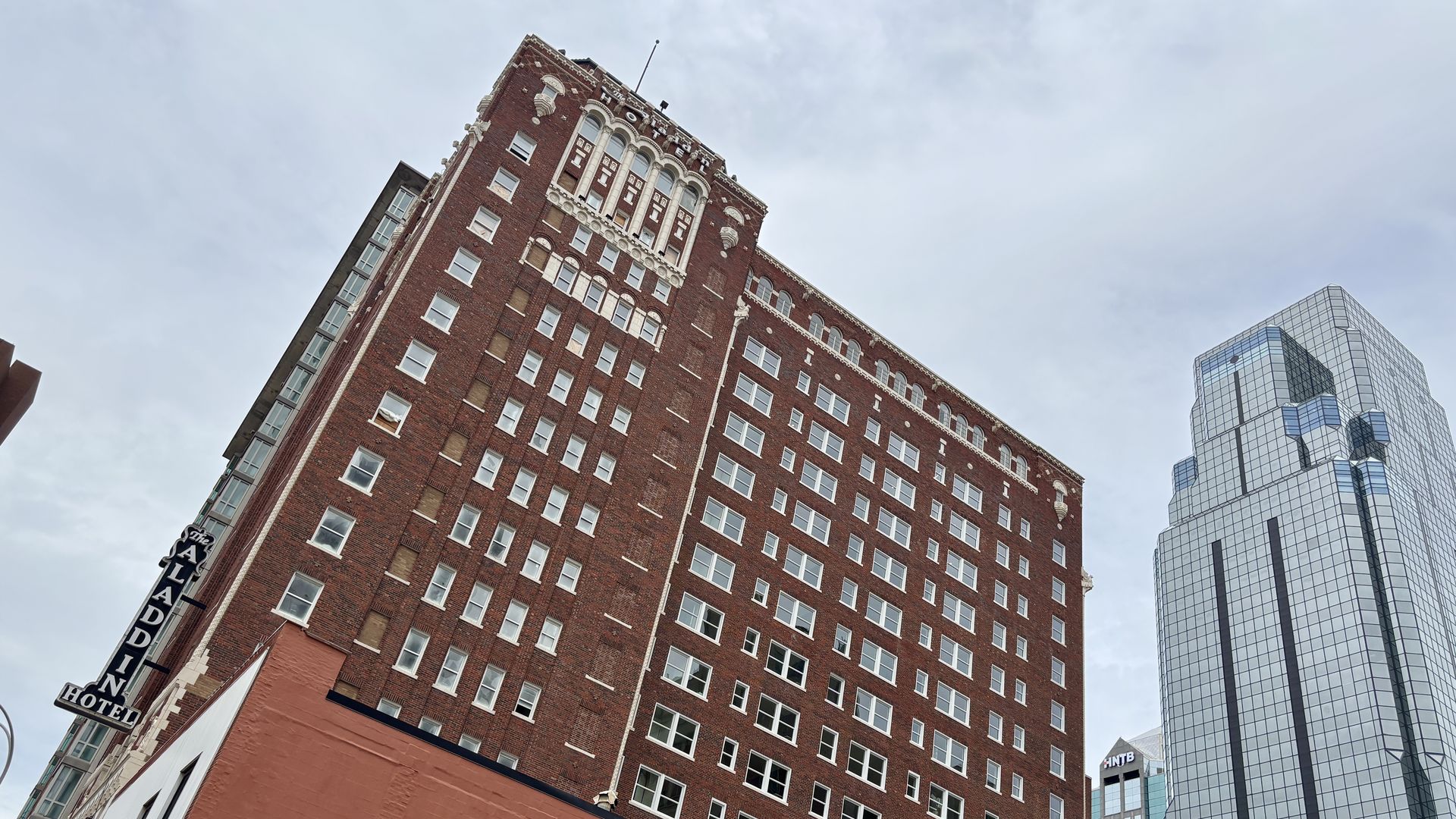 Tilted view of a tall red brick hotel with rows of white-framed windows and decorative white trim at the top, with the words "Aladdin Hotel" visible on the side and at the top, beside a modern glass office building under a blue sky.
