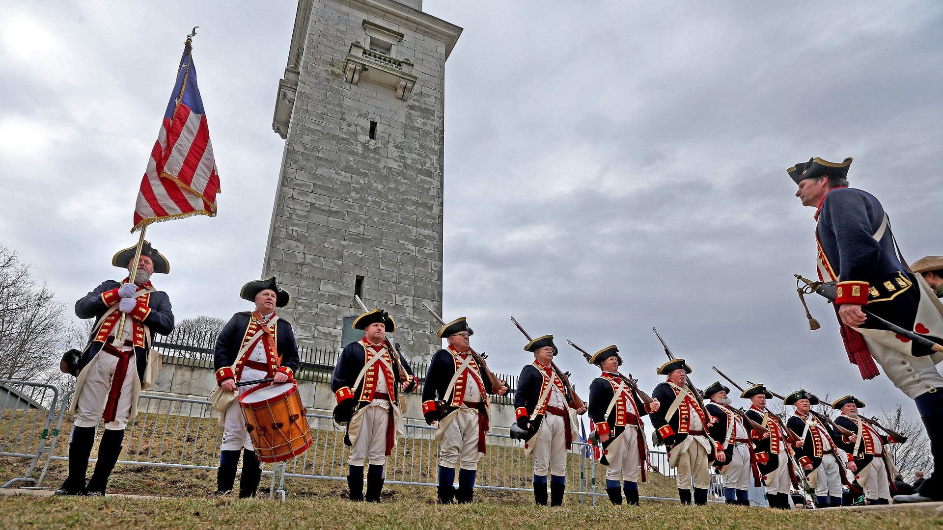 Line of Revolutionary War reenactors in blue coats, white pants, and red trim stand before a tall stone tower; flag bearer with the American flag, drummer at left, cloudy sky above.