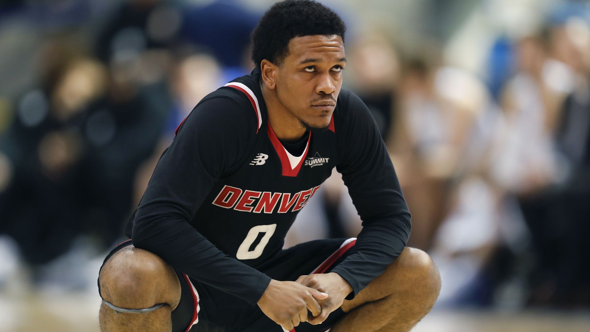 A basketball player crouches down with a pensive facial expression while wearing a black jersey with the word Denver on it.