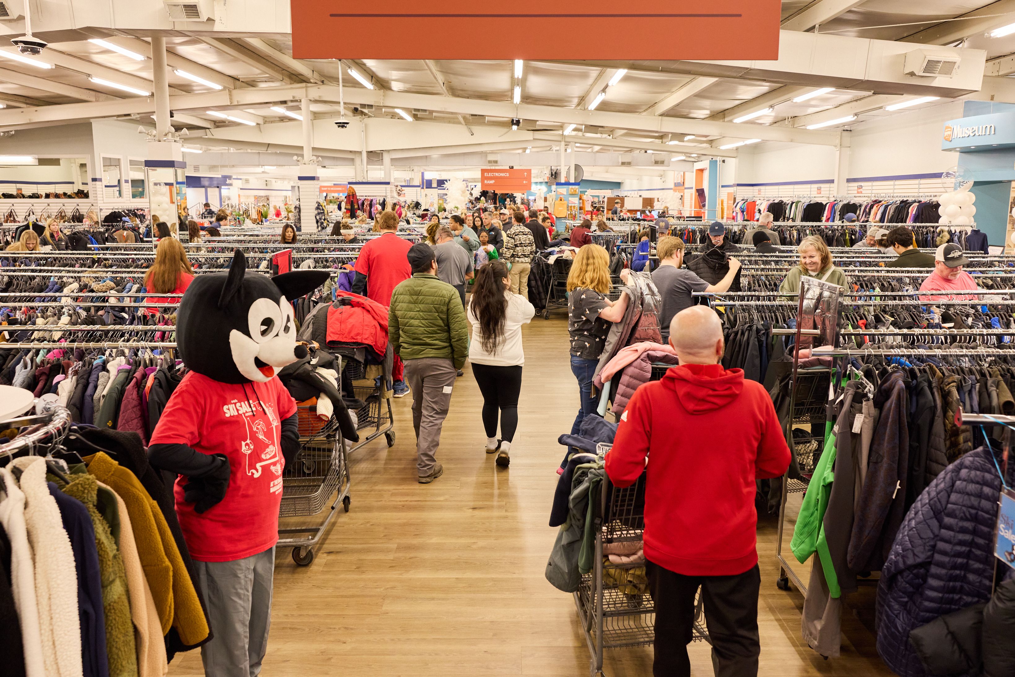 Busy thrift store crowded with clothing racks and shoppers. A Mickey Mouse mascot in a red shirt stands in the foreground; orange overhead sign and bright fluorescent lighting illuminate the scene.