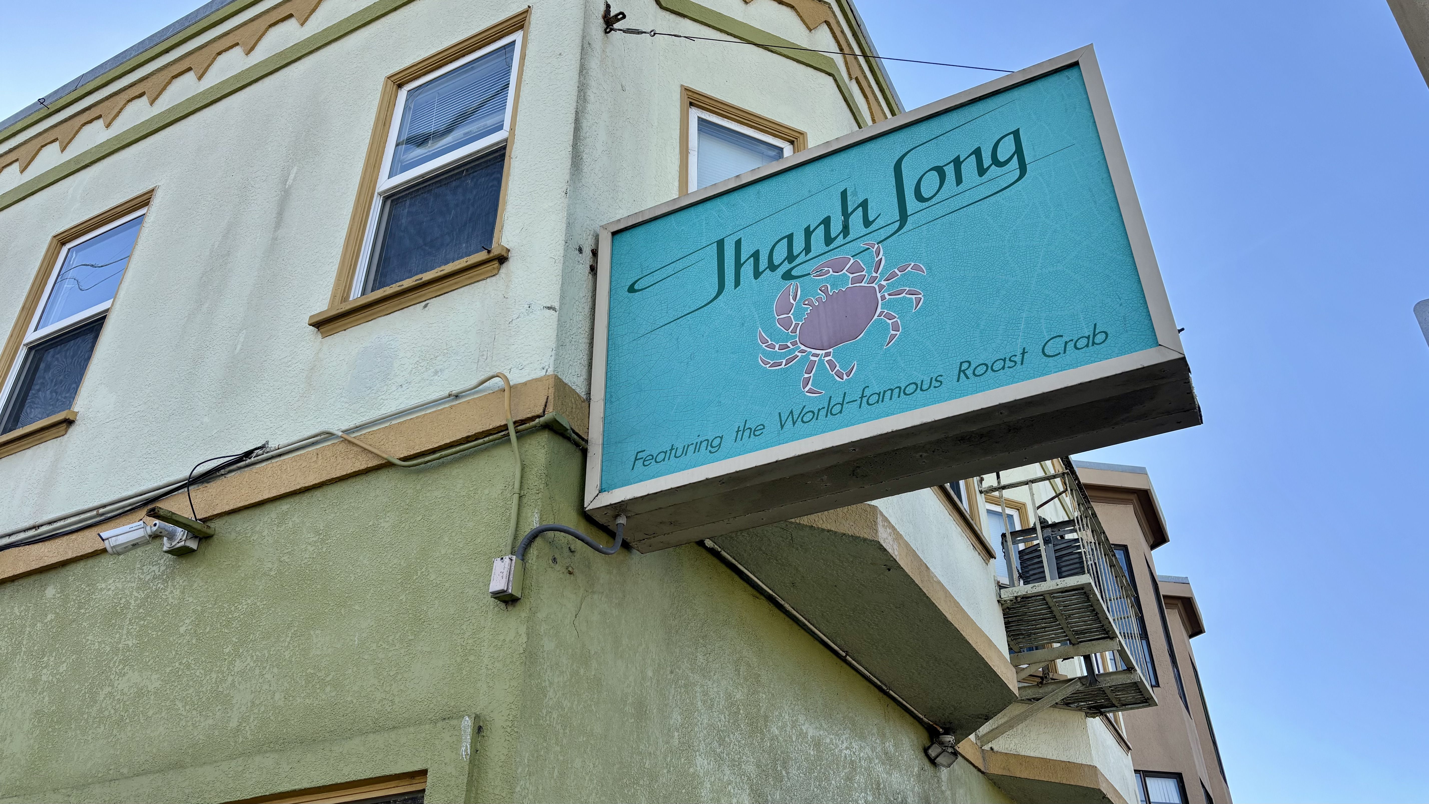 Turquoise restaurant sign with a pink crab and text "Thanh Long Featuring the World-famous Roast Crab" on a beige and green building under clear blue sky.