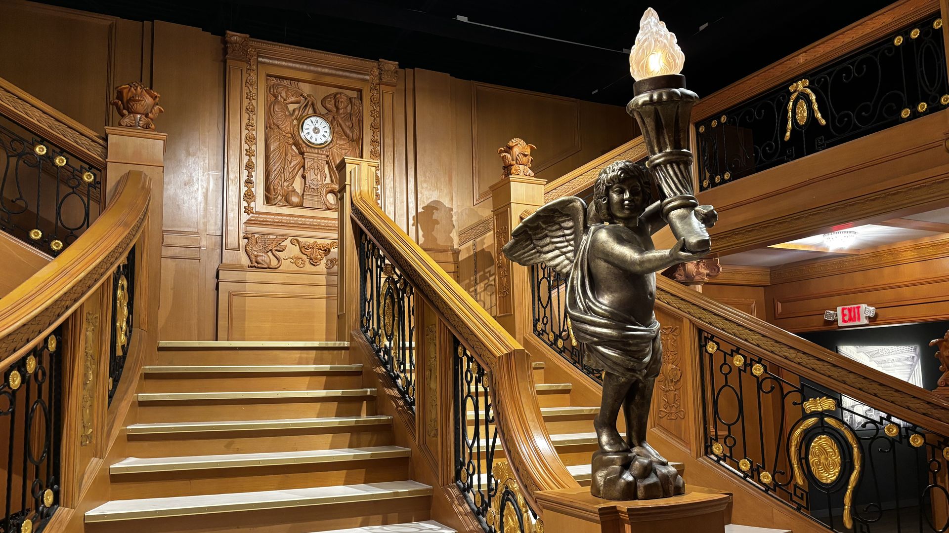 Wooden grand staircase with black and gold ornamental railings, a bronze cherub statue holding a torch, and a wooden wall clock with carved figures above the stairs.