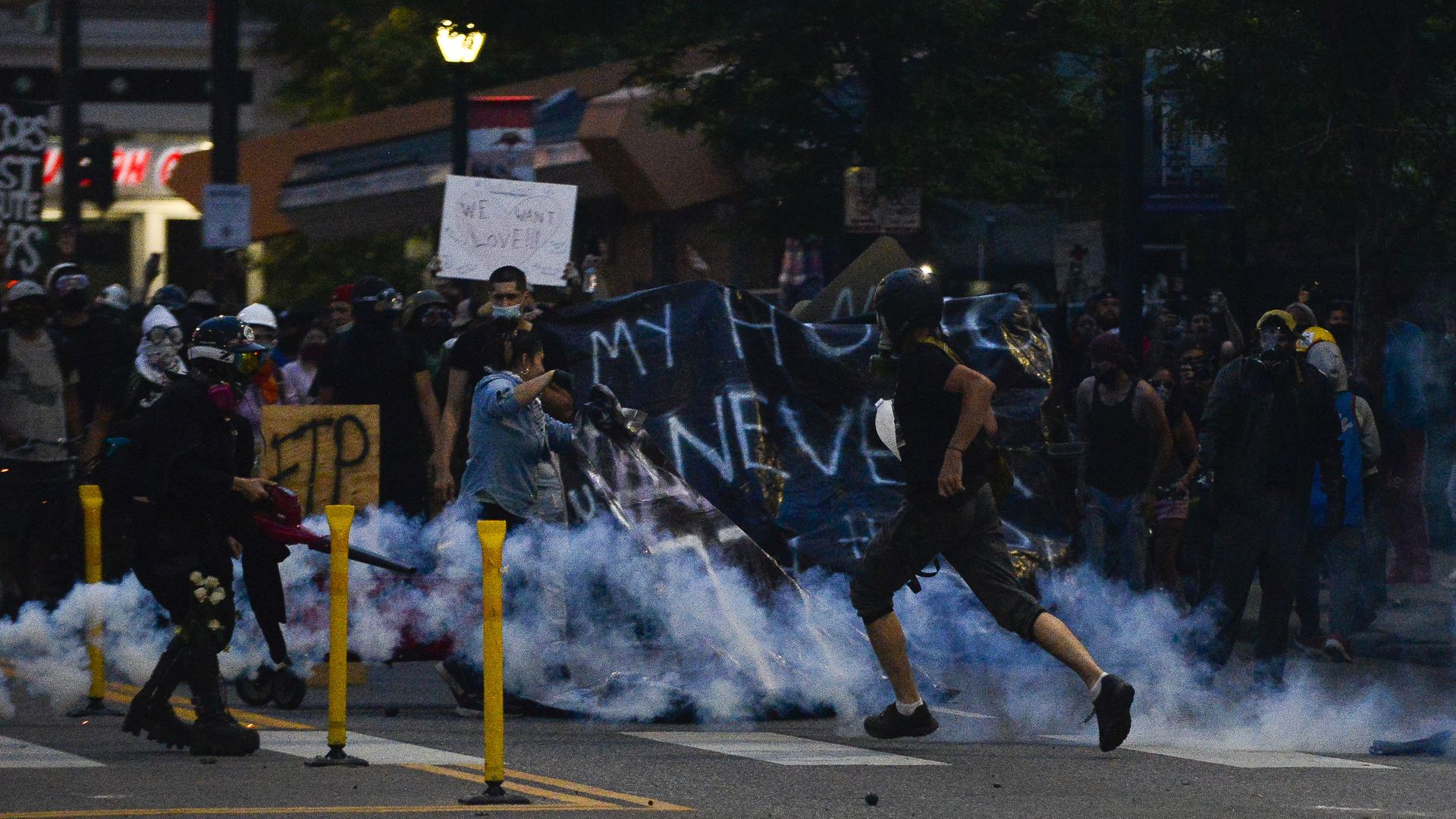 Nighttime street protest with a crowded group of masked protesters and police in helmets. Smoke on the pavement, banners and signs, and lit street lamps in the background.