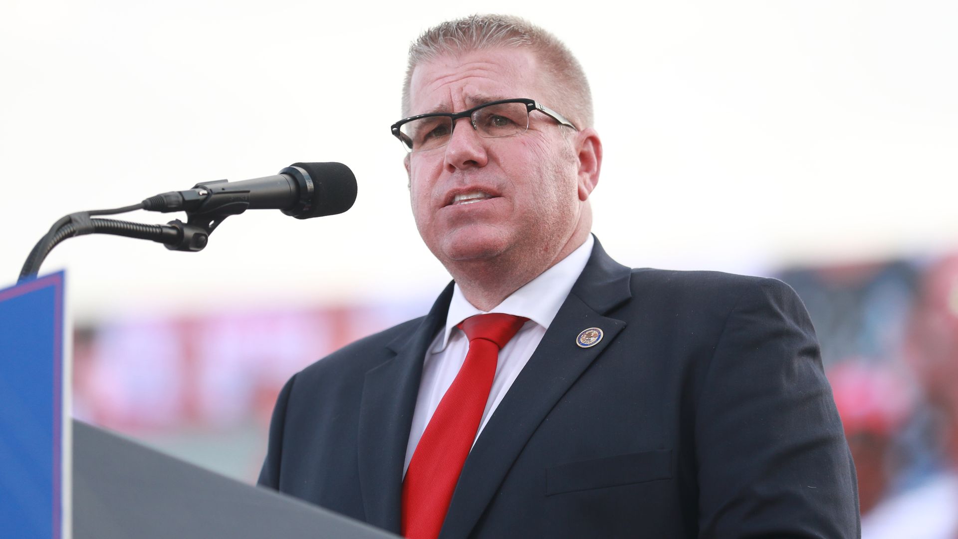 A middle-aged man in a dark suit, white shirt, and red tie speaks into a microphone at a podium; he wears glasses and a circular lapel pin, with a blurred crowd in the background outdoors.