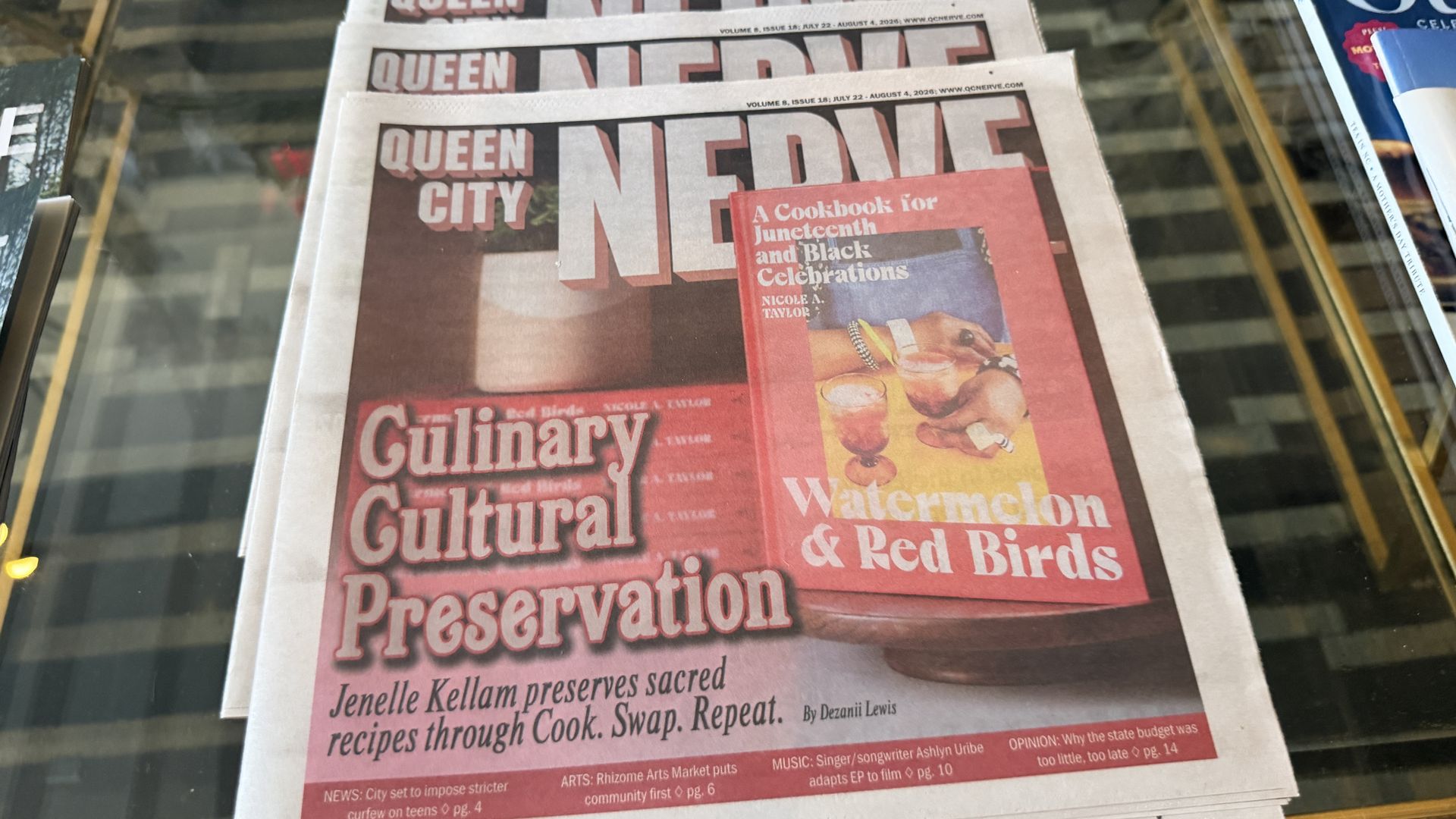 Stack of Queen newspaper front pages on a glass table, bold white and red headlines. The visible cover mentions Juneteenth and Black celebrations, culinary culture, and preservation.