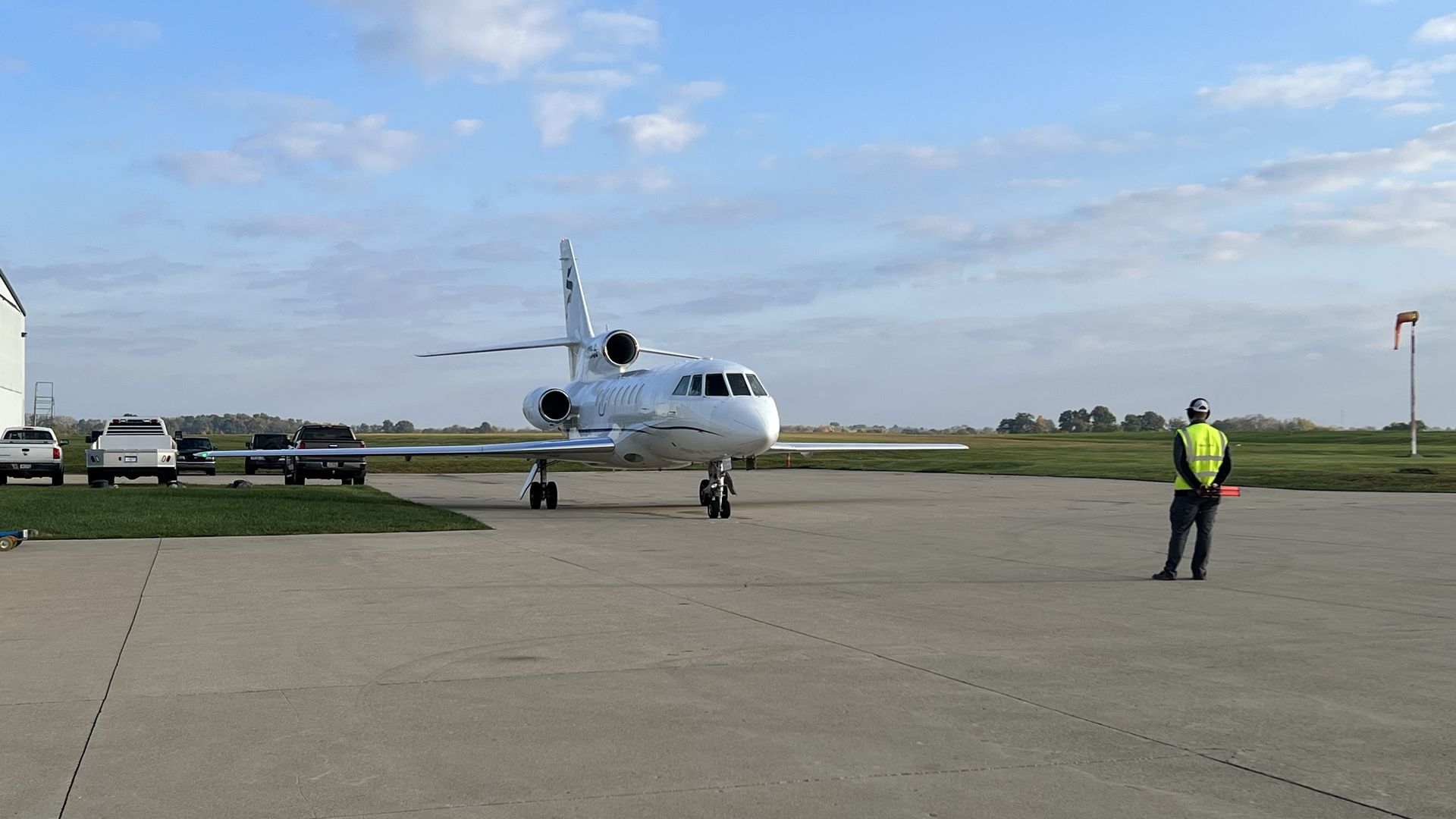 White twin-engine business jet on a runway with rear-mounted engines. A person in a neon safety vest stands to the right; several cars on the left and a windsock in the distance under a blue sky.