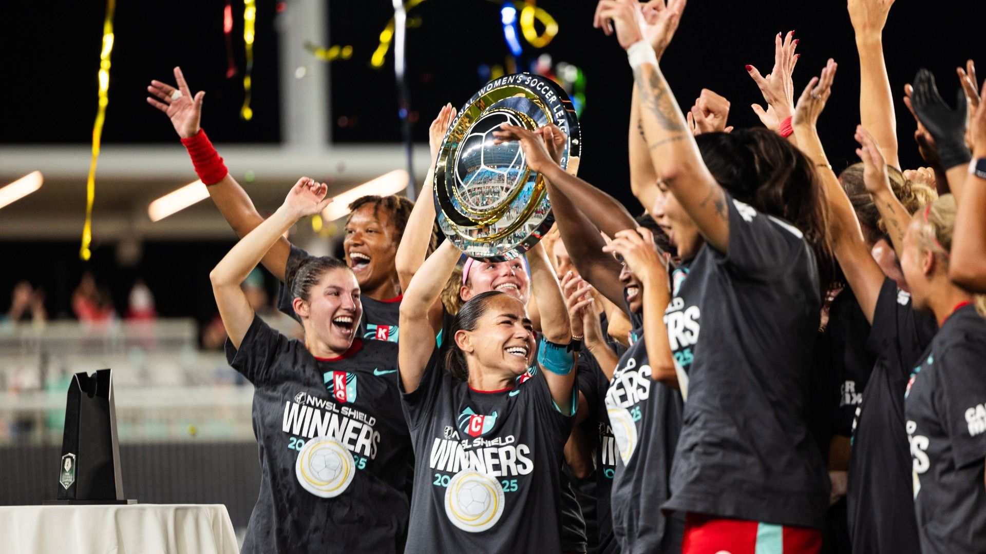 KC Current players in black shirts saying "winners" celebrate by holding up the NWSL Shield with streamers in the background.