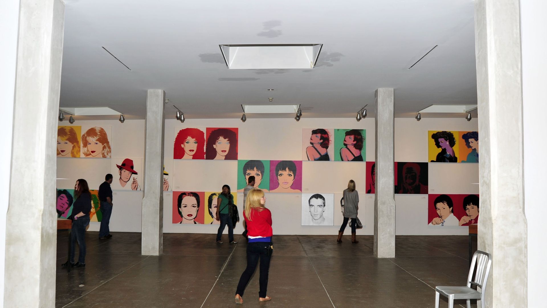 People viewing colorful pop art portraits of various faces on white walls in a gallery with concrete pillars and floor, under ceiling lights.