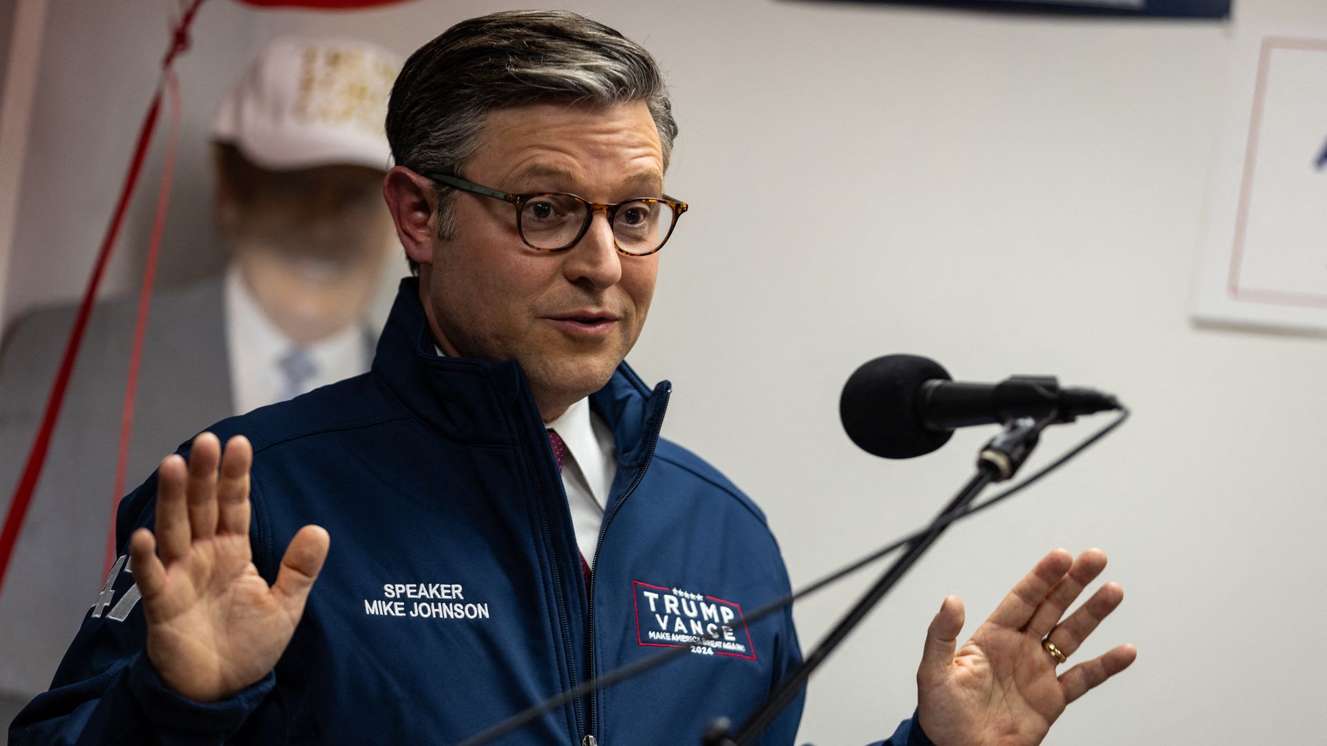 Mike Johnson, wearing a blue windbreaker and speaking at a microphone in front of a white wall and a Trump cut-out.
