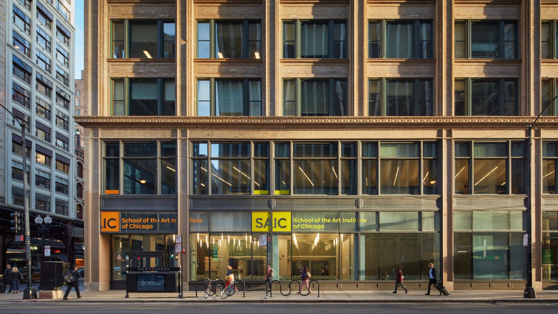 Street view of a brick building with large windows and signs reading "School of the Art Institute of Chicago" in orange and yellow panels, with pedestrians and bicycles outside.