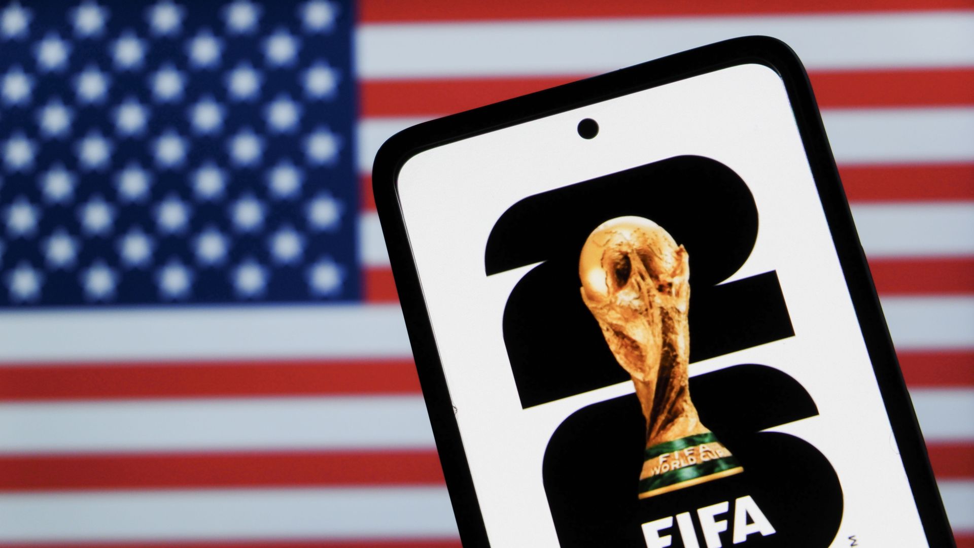 Tilted smartphone displays the FIFA app icon with a golden World Cup trophy; a blurred American flag fills the background.