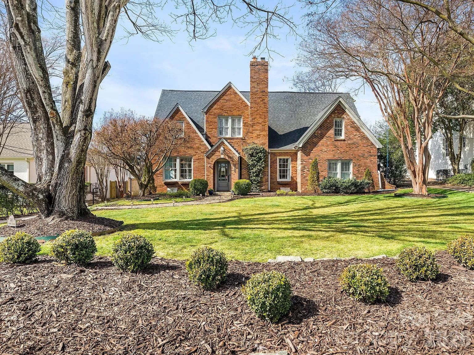 Brick house with gray roof and white-trimmed windows, large front chimney, green lawn, and trimmed bushes with tall trees in front under a clear blue sky.