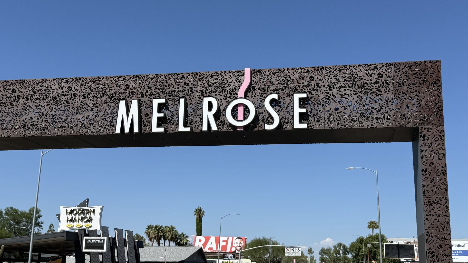 Clear blue sky above a decorative Melrose arch sign with white letters outlined in black, and a pink curved accent. Palm trees and storefronts line the street below.