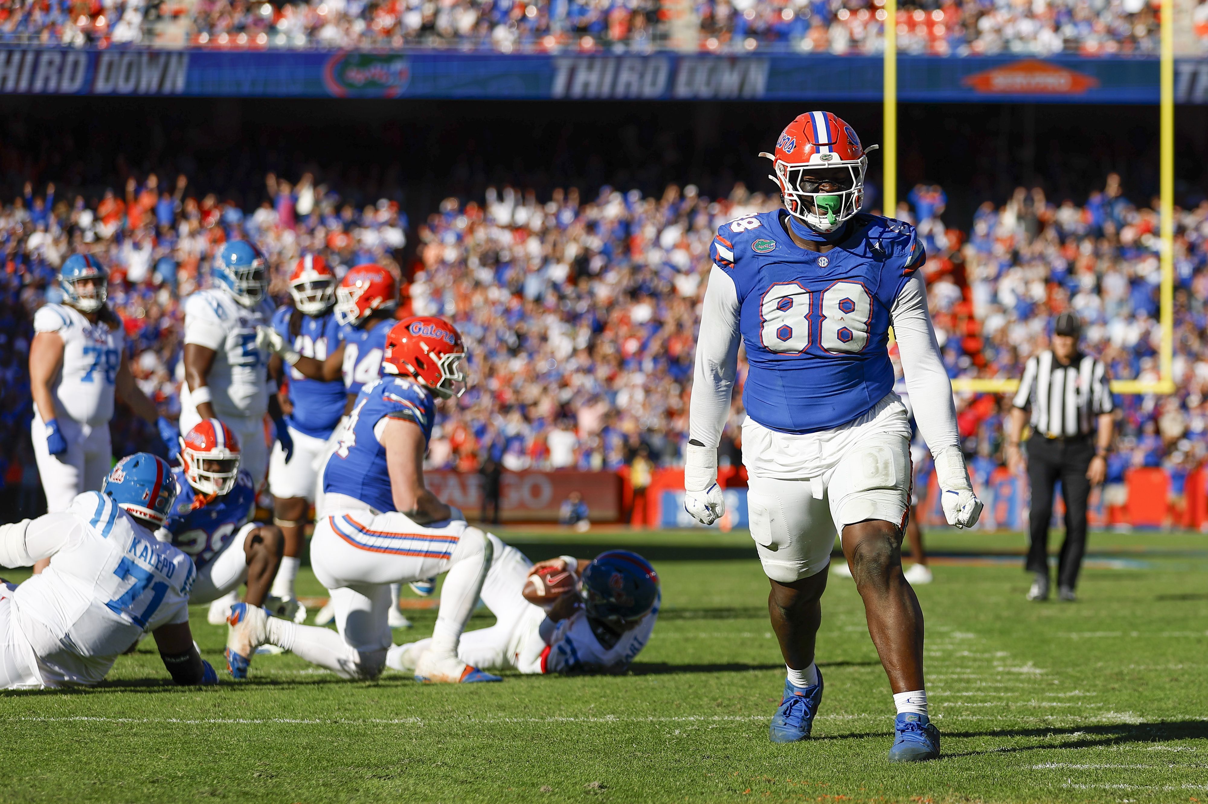 Florida Gators football player #88 in blue jersey and orange helmet walks across the field as teammates and opponents lie on the turf after a play; a crowded stadium is in the background.