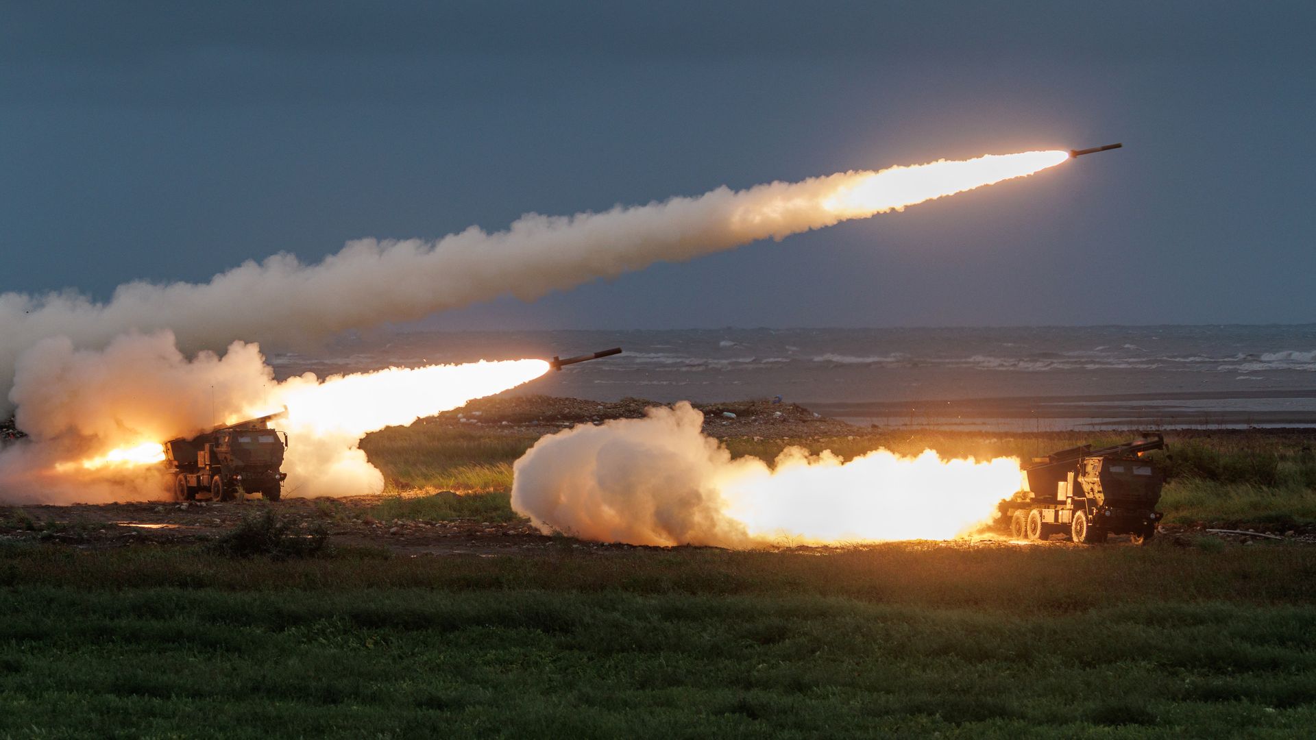 Multiple missiles launch from ground-based launchers across a barren landscape at dusk. Fiery exhaust trails and billowing white smoke arc across a deep blue sky with distant hills.