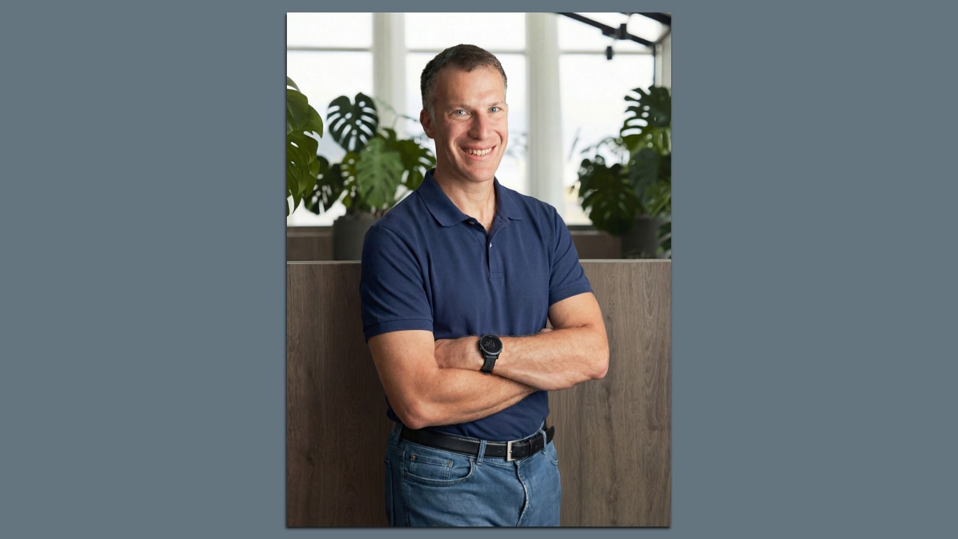 Smiling man with short hair in a navy polo shirt and jeans stands with arms crossed in an office space, with large leafy plants and bright windows in the background.