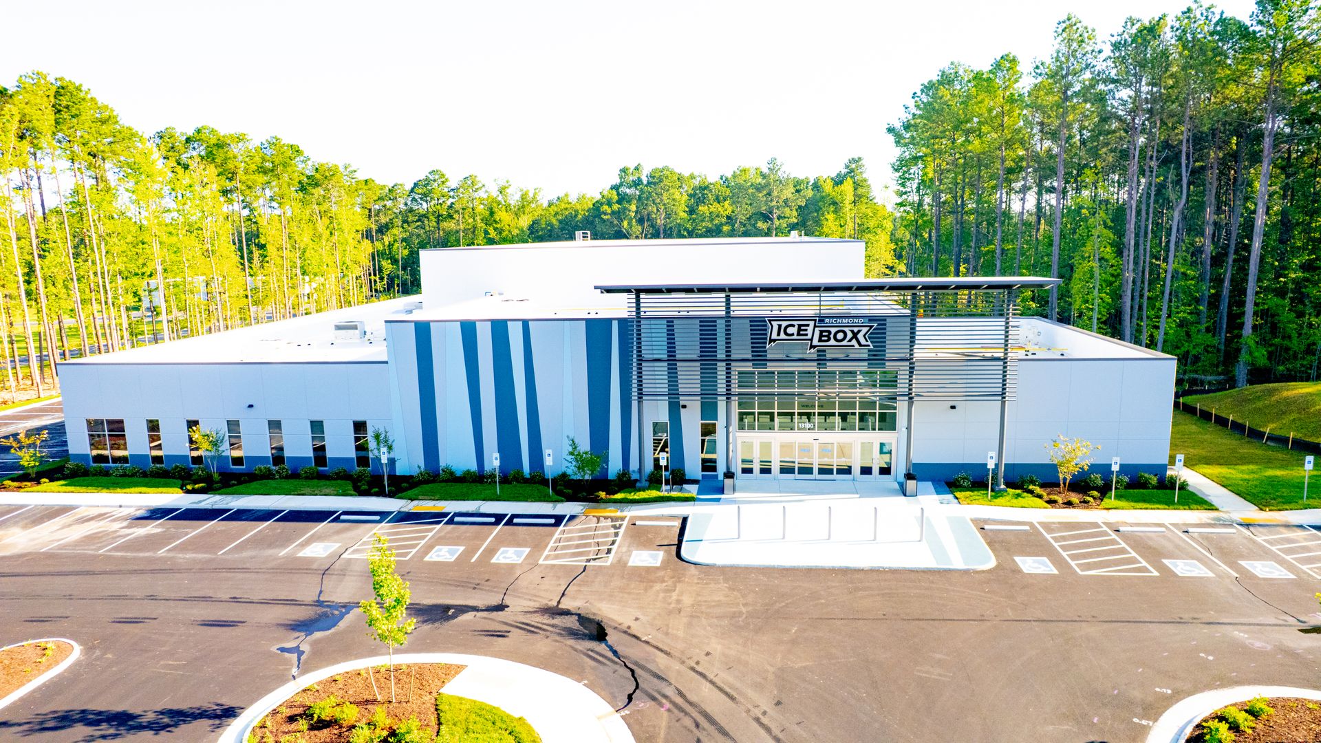 A modern white commercial building with blue vertical panels and a prominent "ICE BOX" sign, in a tree-lined parking lot with a dense forest behind it.
