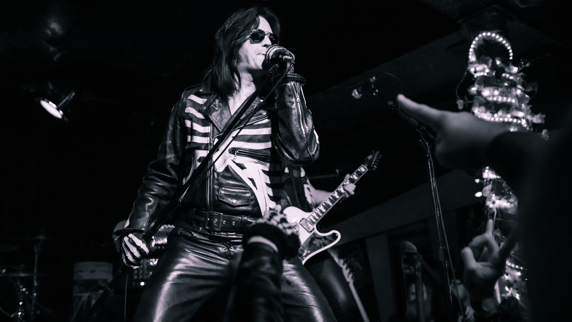 Black and white photo of a rock singer in a leather skeleton jacket and sunglasses singing into a microphone on stage, with guitarist and fans' hands pointing in foreground.