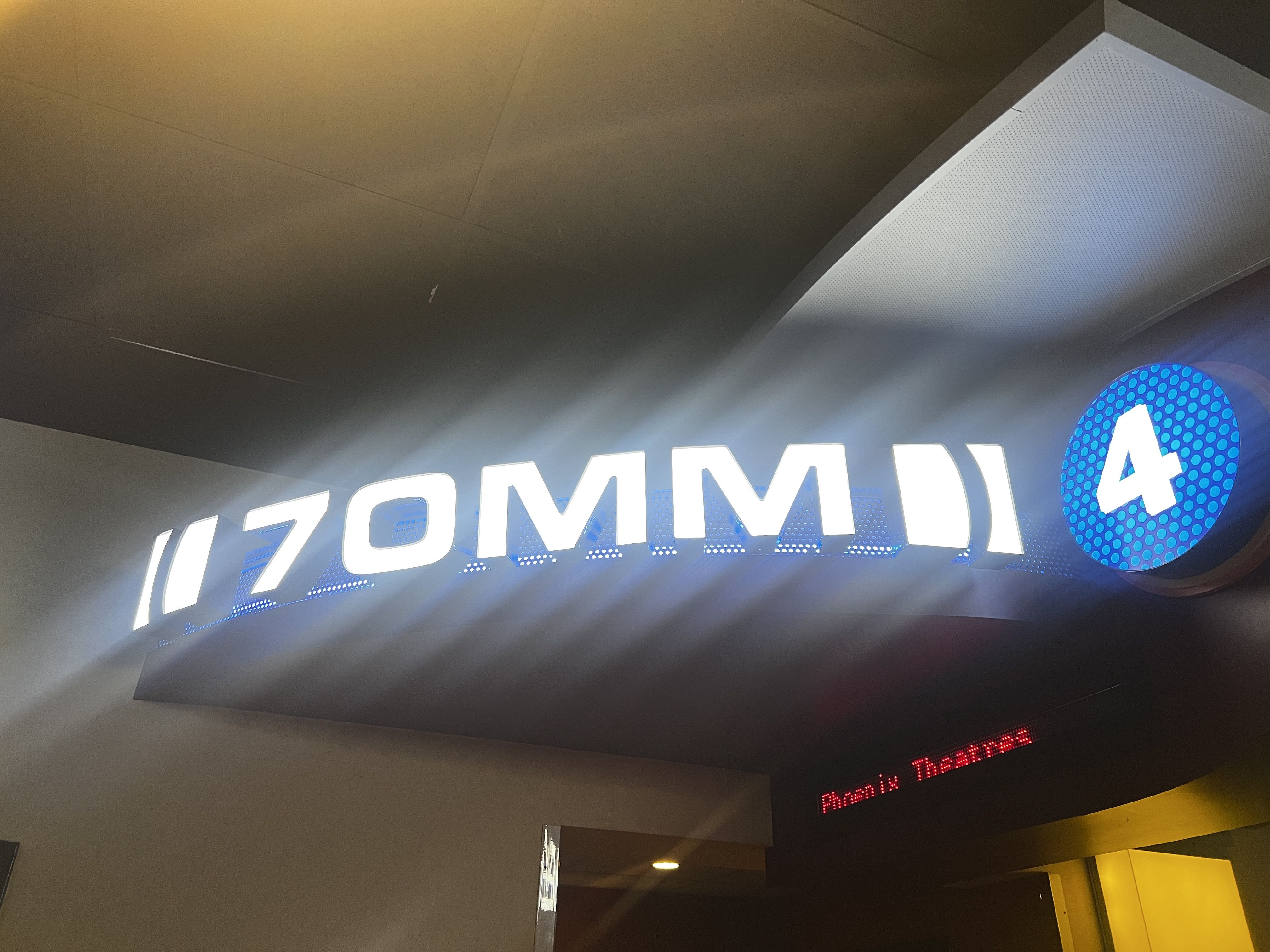Interior of a cinema lobby with a slanted ceiling. A large illuminated sign with bold white letters spans a wall, featuring a blue circular logo with a white A; a red LED marquee glows to the right.