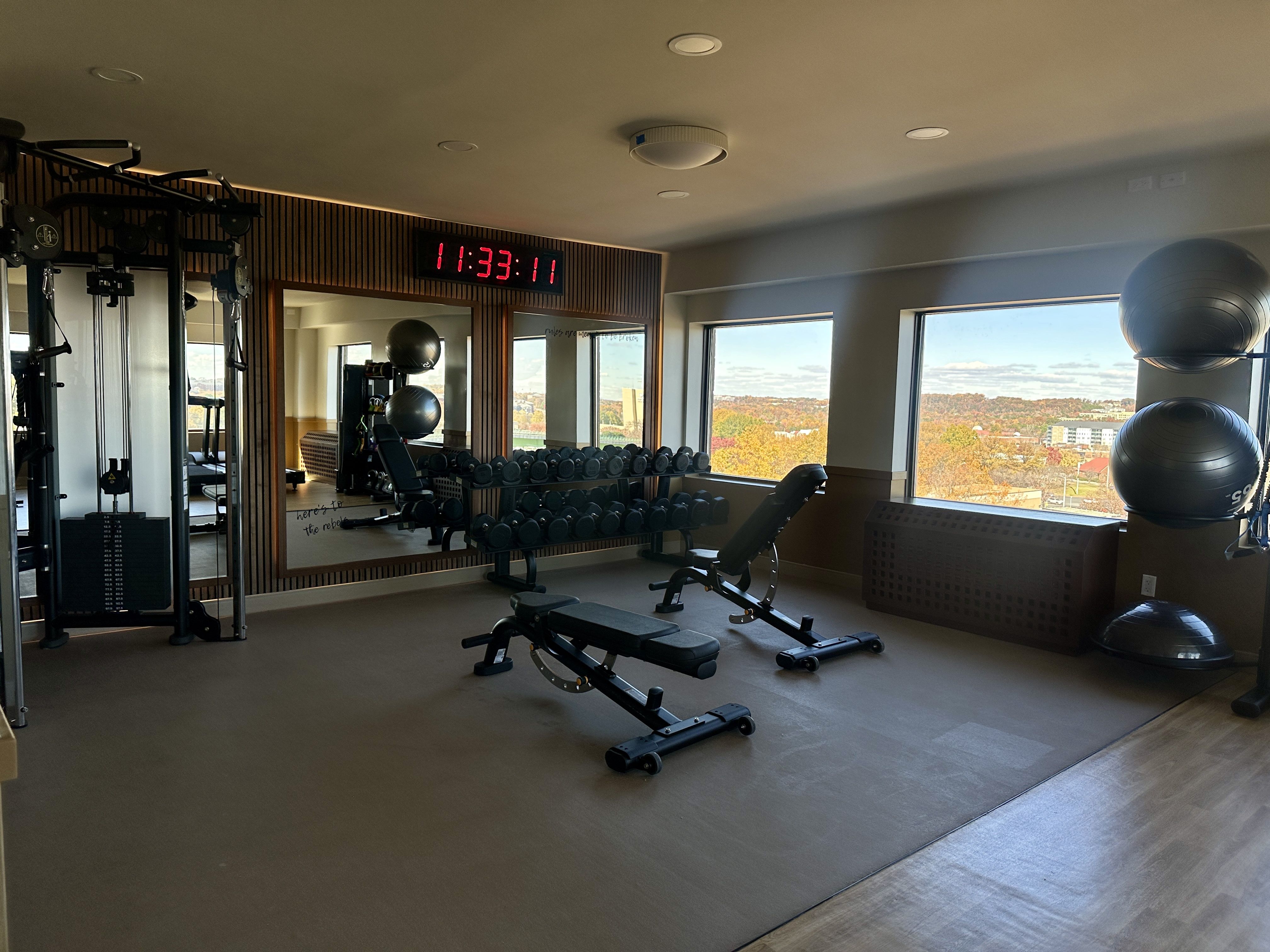 Bright gym room with two black workout benches, dumbbell rack in front of large mirror showing reflections, a cable machine, stacked gray exercise balls, and windows with an autumn outdoor view.