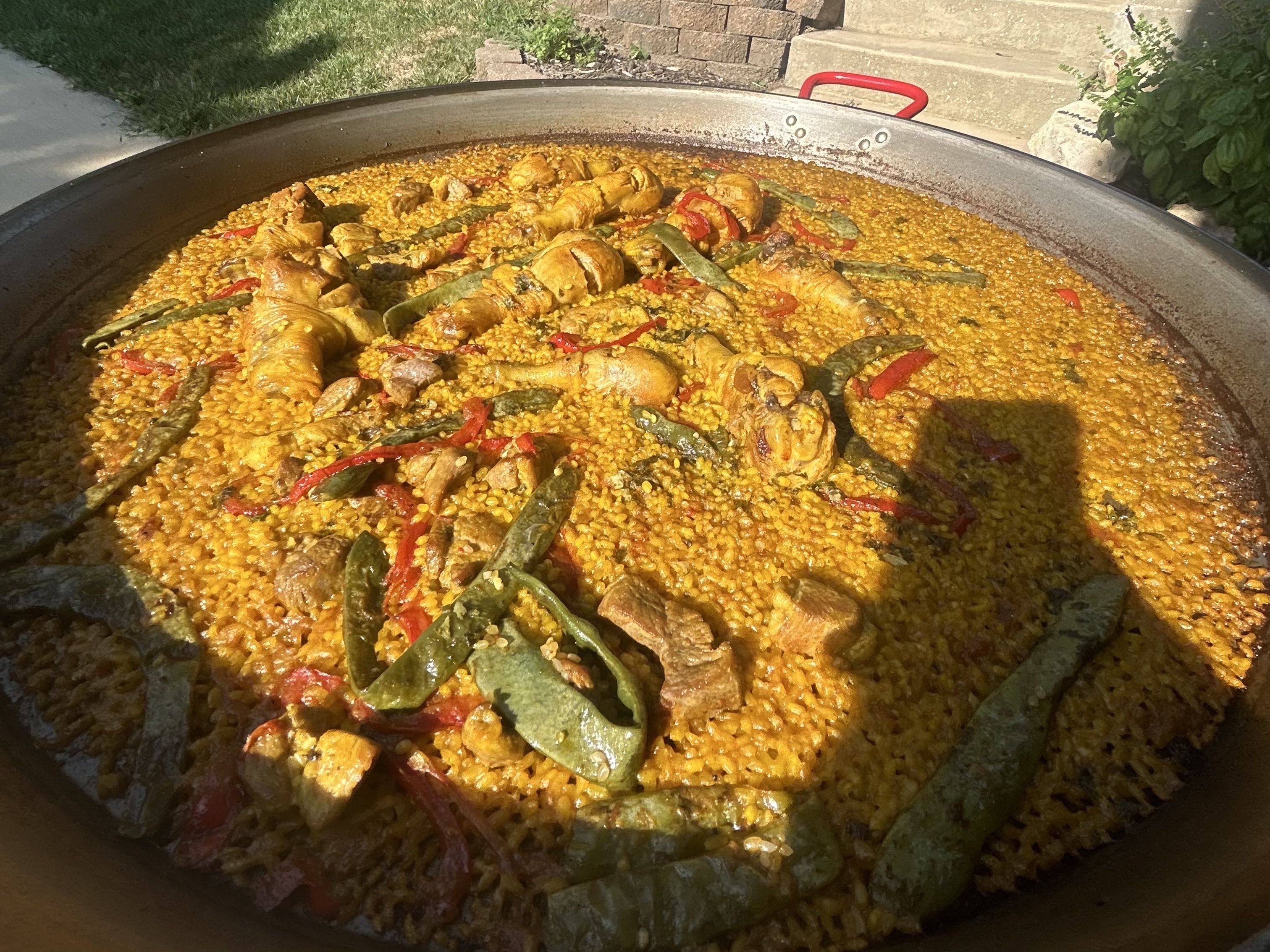 Large pan of homemade paella with yellow rice, chicken drumsticks, red bell peppers, and green beans cooked outdoors in sunlight.