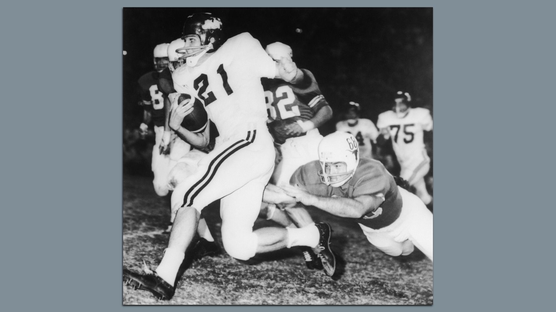A black-and-white photo shows a football player carrying the ball while being tackled during a game, with other players in the background.