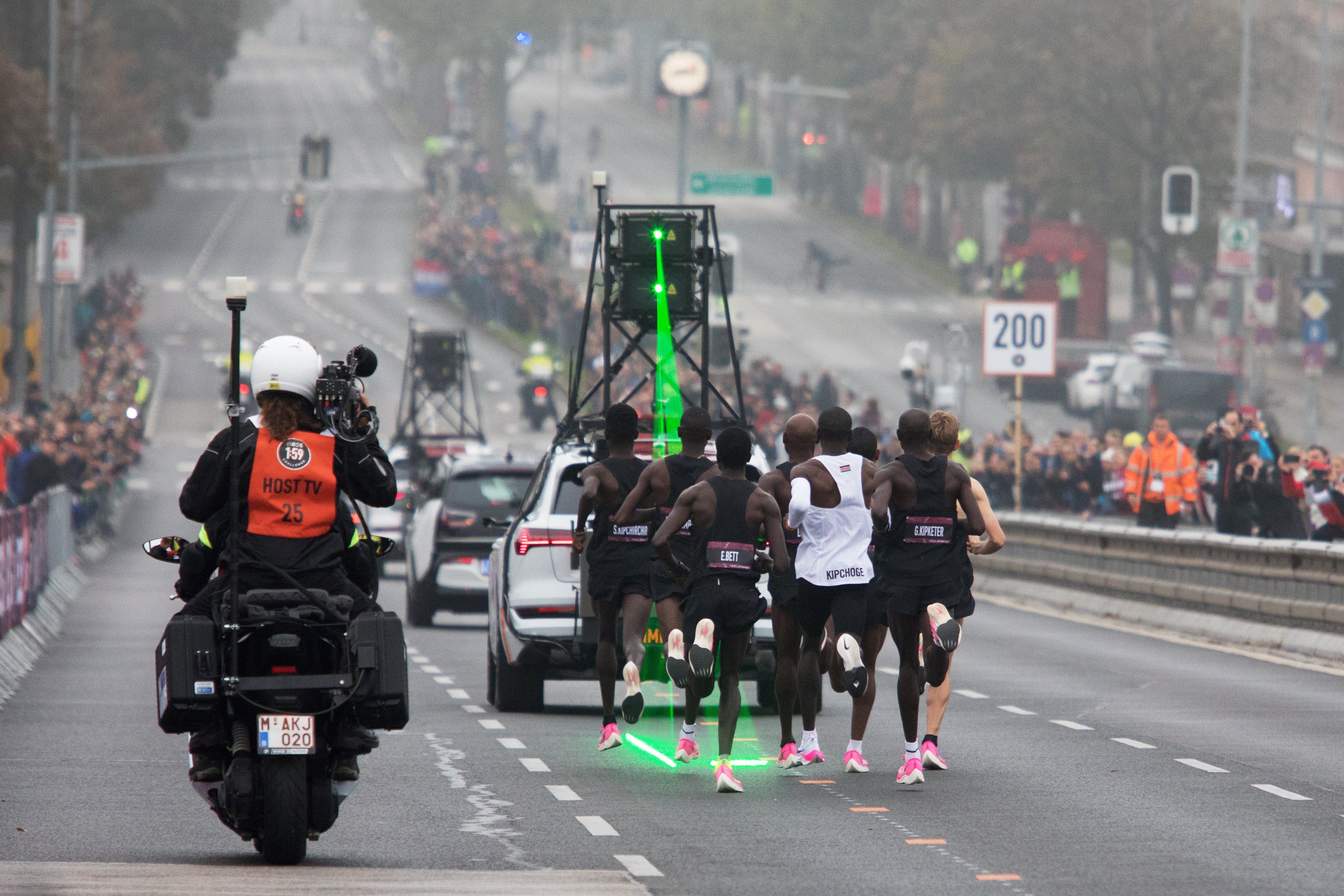 In this image, a group of runners run in close formation behind a car that shines a green laser onto the pavement.