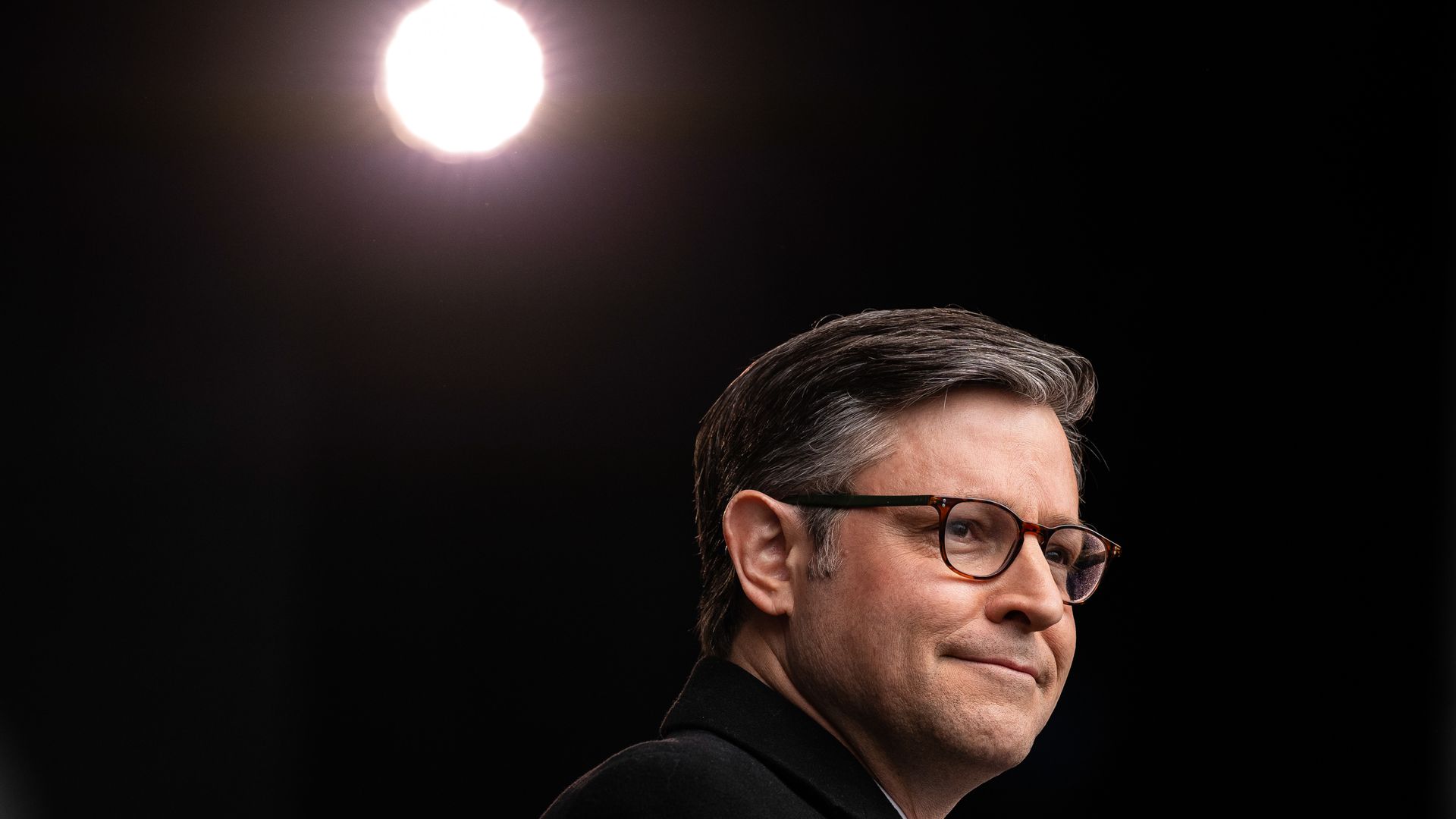 Profile of a man with gray hair and glasses wearing a black jacket, smiling slightly against a dark background with a bright, round light above him.