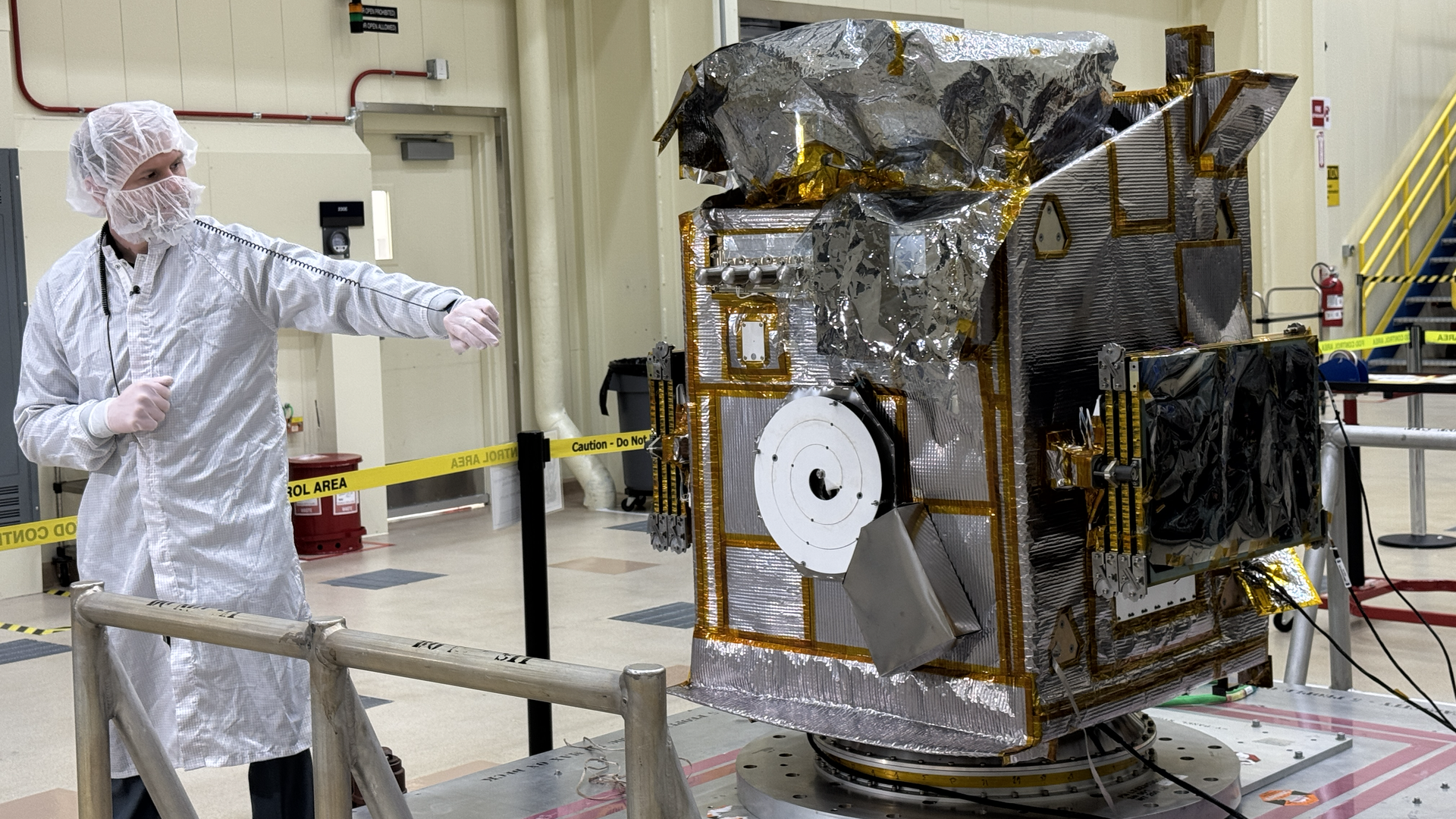 A man in a white coat and facemask points toward a large metal satellite. 