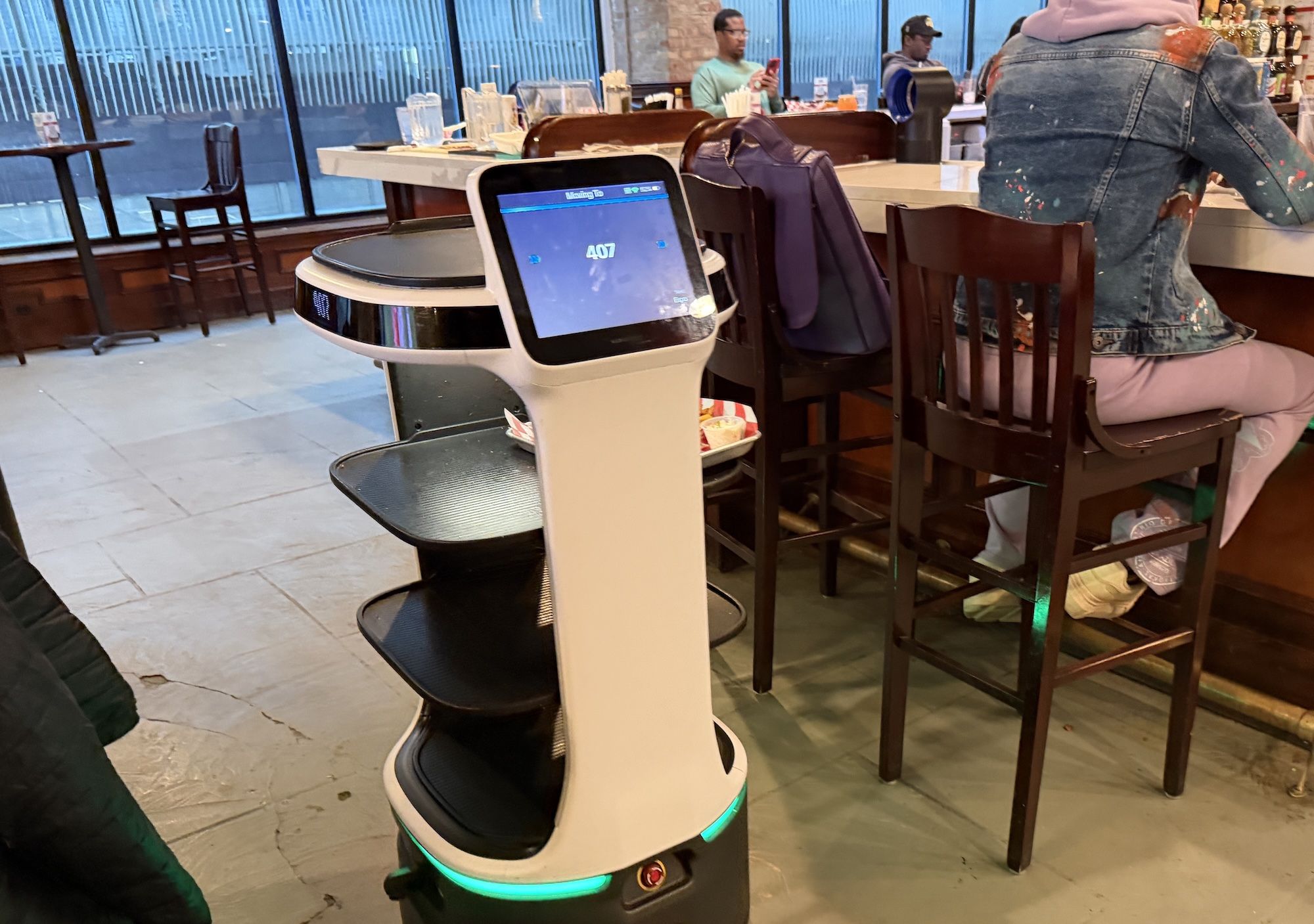 White autonomous service robot with a touchscreen displaying 407 stands at a bar in a casual restaurant; a person in a paint-splattered denim jacket sits on a stool at the counter.