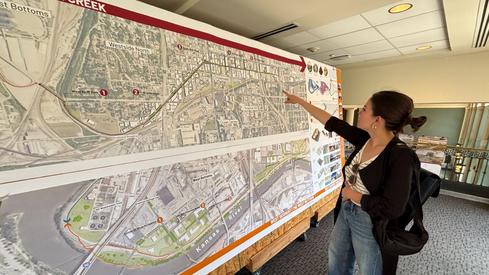 Woman with a backpack points at a large city map mounted on a wall in a hallway; the map shows streets, a river, numbered markers, and a red border along the top.
