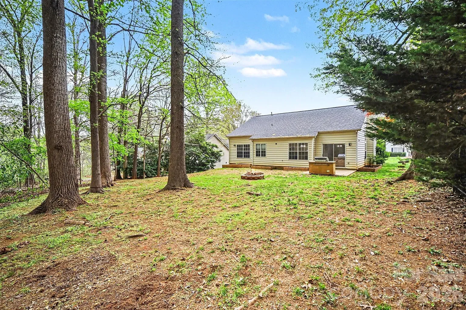 Backyard of a single-story beige house with a gray roof. A wooden deck with a grill sits at the back; a sliding glass door is visible. A circular fire pit rests on the green lawn, with tall trees framing the yard.