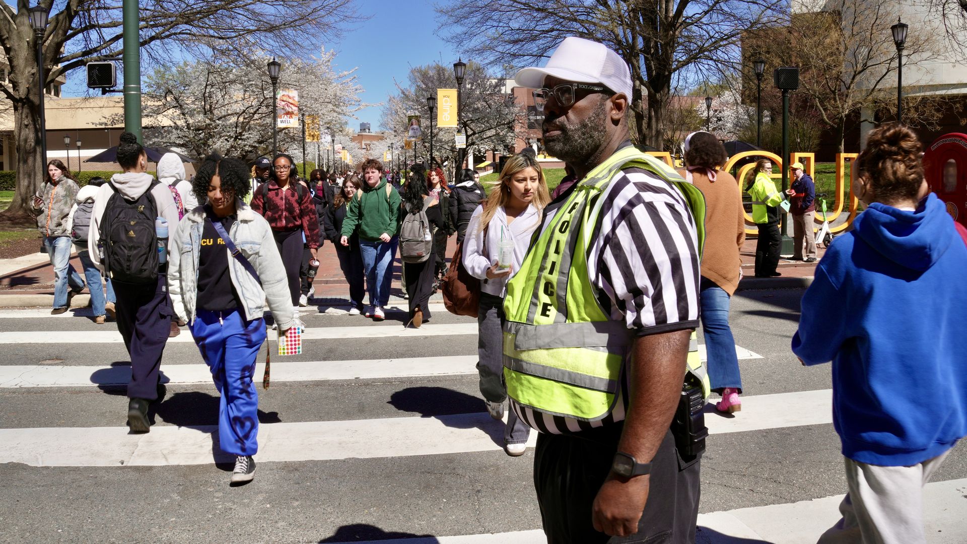 Students cross a sunny campus crosswalk as a security officer in a neon vest and striped shirt stands in the foreground; blooming trees and campus buildings in the background.