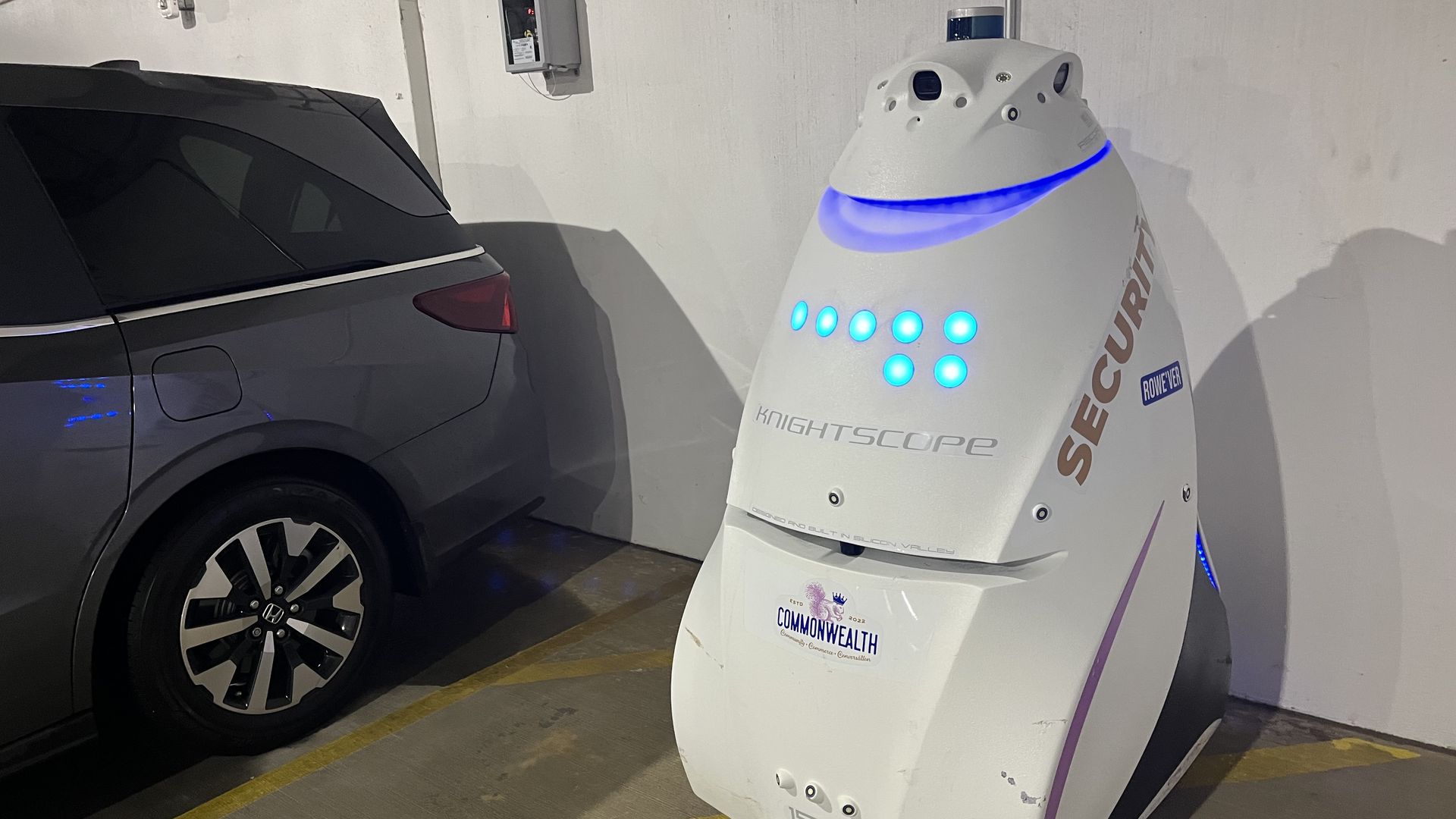 White Knightscope autonomous security robot with glowing blue lights parked next to a dark gray Honda vehicle in a parking garage with concrete floor and white walls.