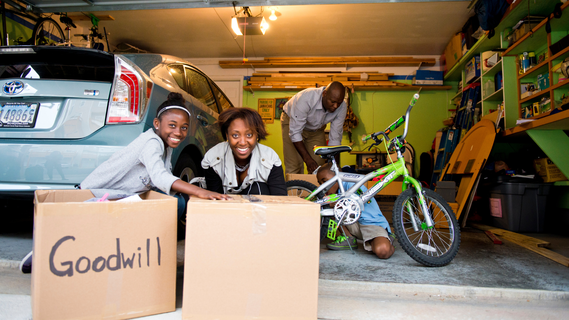 Three people are in a garage. One man is fixing a bike. In front of him, two women are getting packing boxes ready.