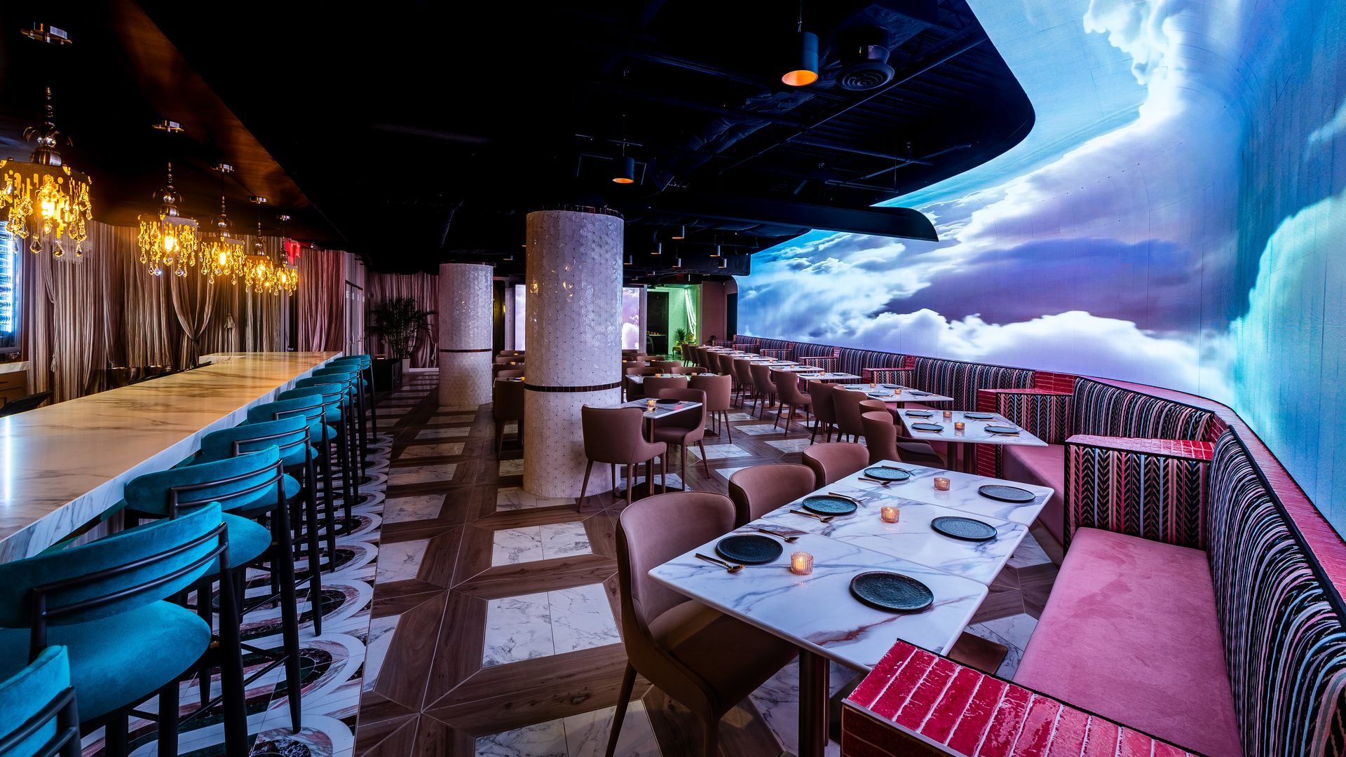 Luxurious restaurant interior with a long marble bar and turquoise stools on the left, gold chandeliers, marble columns, and pink striped booths; a large blue sky mural spans the right wall.