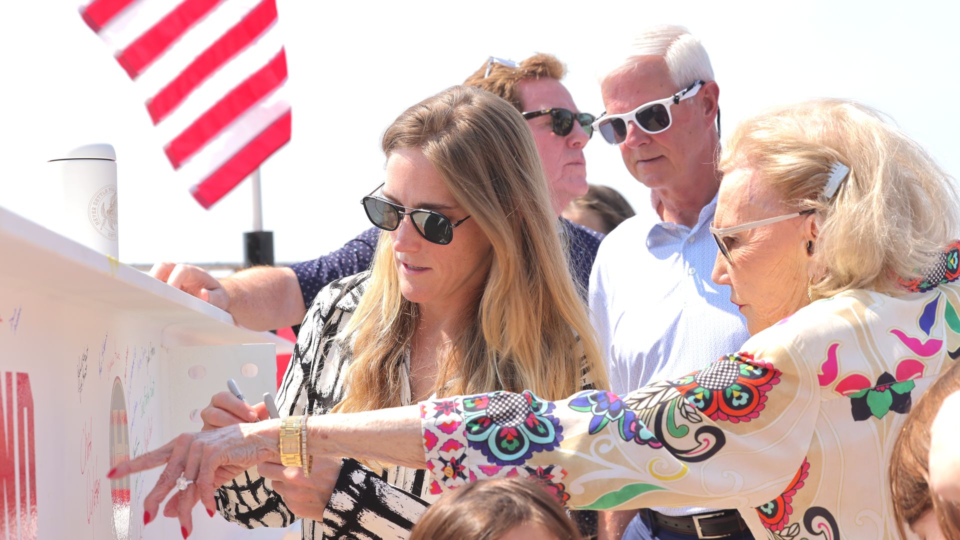 ChatGPT said: A group of people sign a steel beam at a ceremonial event, with an American flag waving in the background under bright sunlight.