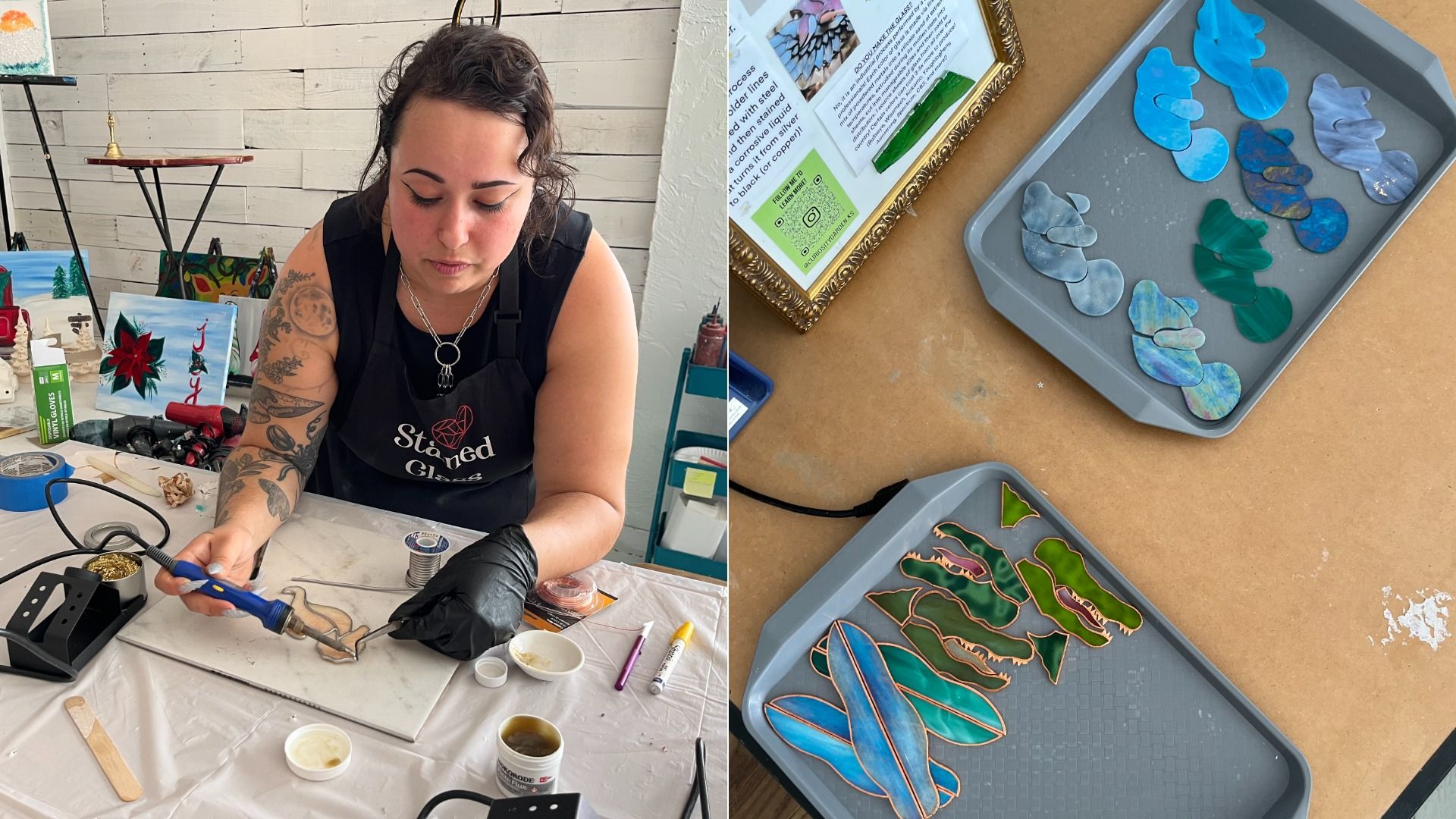 Woman with tattoos and black gloves soldering stained glass pieces at a worktable with tools, paints, and art in a bright room; two trays with colorful glass pieces on a brown surface nearby.