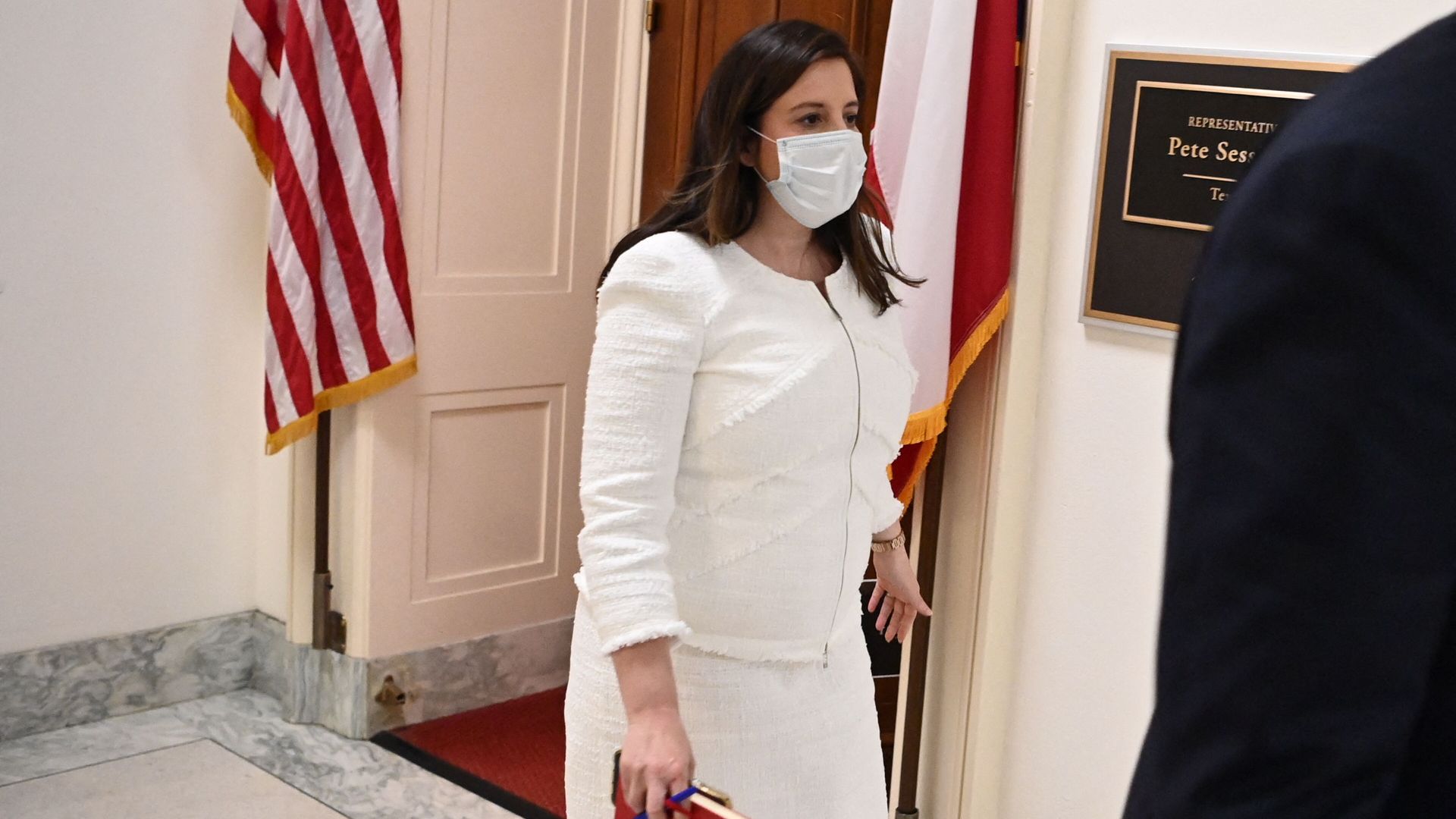 Elise Stefanik walks down a hallway while wearing a mask, with an American flag behind her.