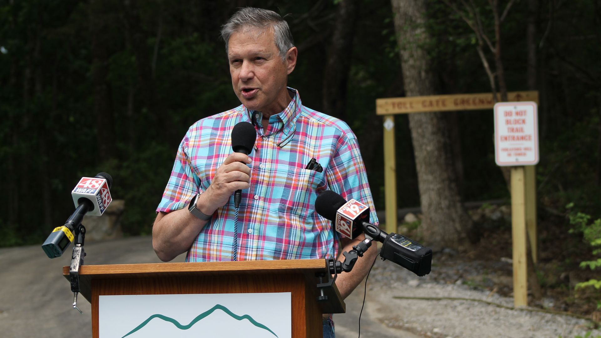 Gray-haired man in a colorful plaid shirt speaks into a microphone at a podium labeled "Land Trust" outdoors, with news microphones and a forest backdrop; a "Do Not Block Trail Entrance" sign is visible.