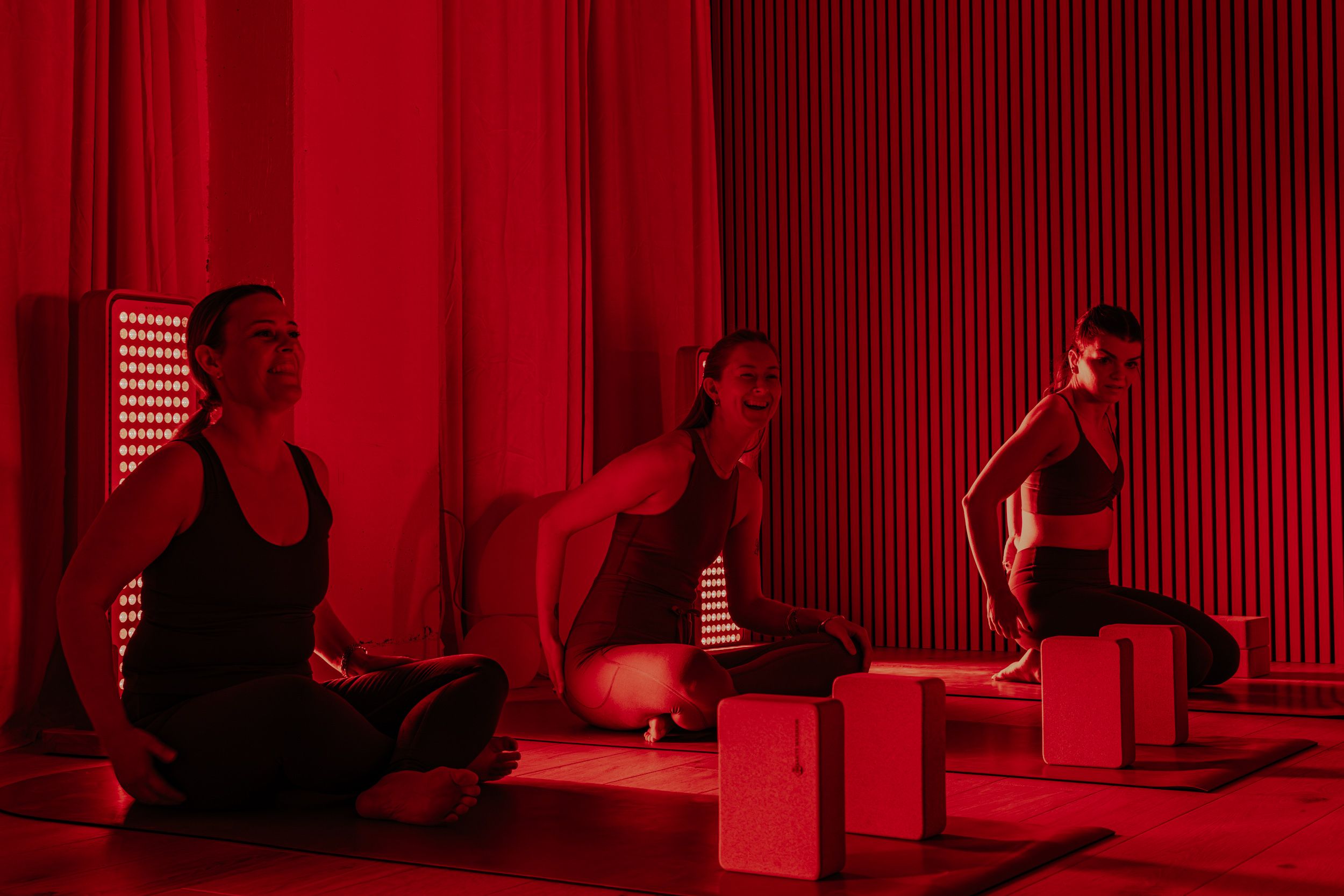 Three women in black workout clothes sitting on mats in a room with red lighting and wooden floor, with yoga blocks in front of them, smiling and stretching.