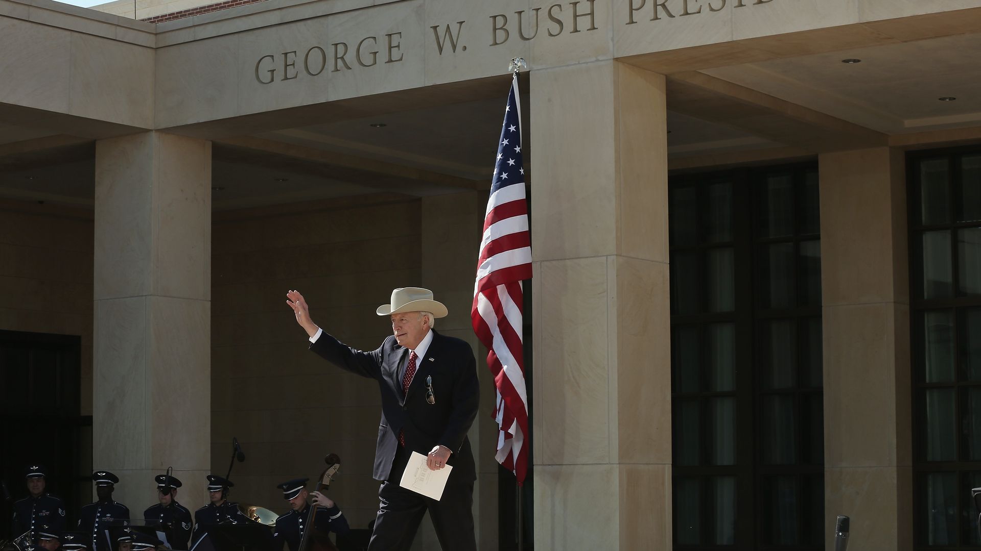 Dick Cheney, wearing a suit and cowboy hat, waves while walking past an American flag at the George W. Bush Presidential Center with a band playing in the background.