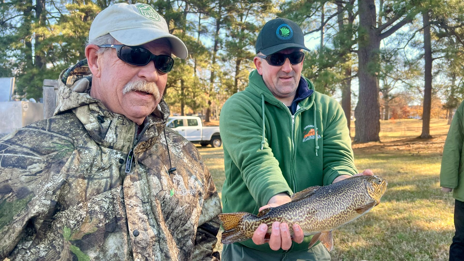 Two men outdoors in a wooded area during daylight, one in camouflage jacket and orange gloves, the other in green hoodie and cap, holding a large brown-patterned fish over a net.