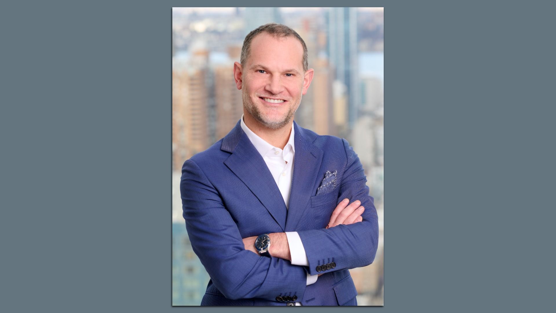 Portrait of Justin Dini in a blue suit and white shirt with a pocket square, arms crossed, city skyline blurred in the background.