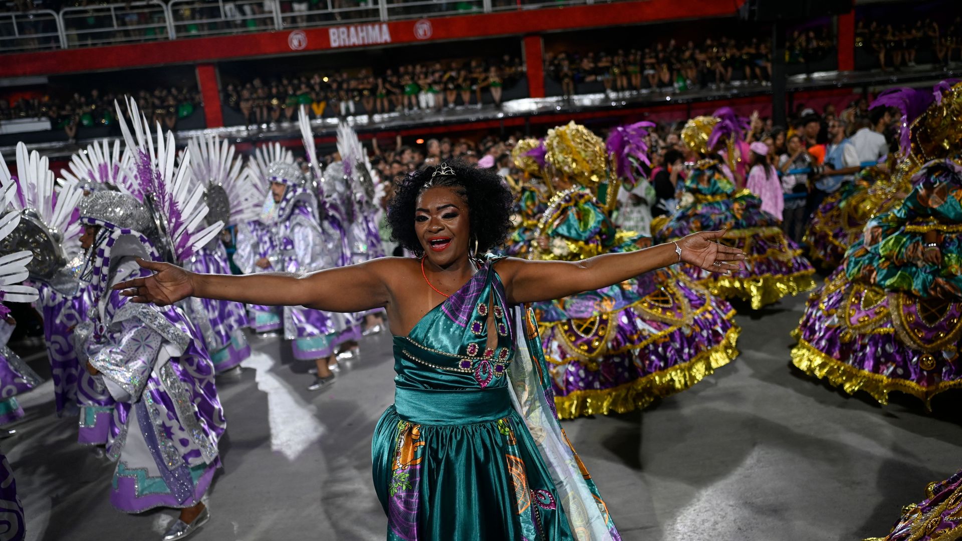 A woman in a birght green and blue dress has her arms outstretched during Carnival in Brazil. She's surrounded by dancers in bright clothing